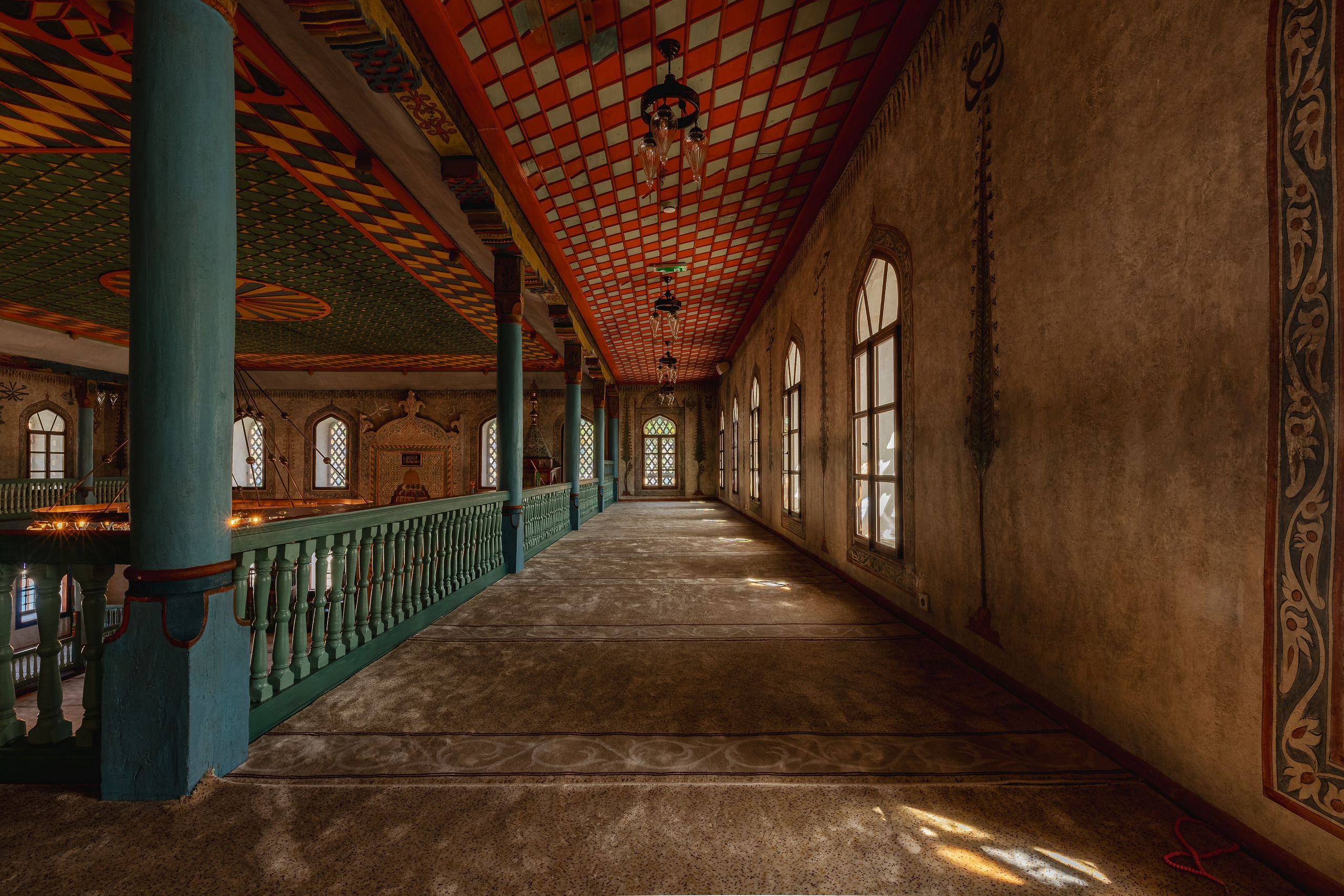 Interior architectural photography of a historic Ottoman mosque featuring geometric ceiling patterns, arched windows, wooden galleries and decorative chandeliers illuminated by natural light.