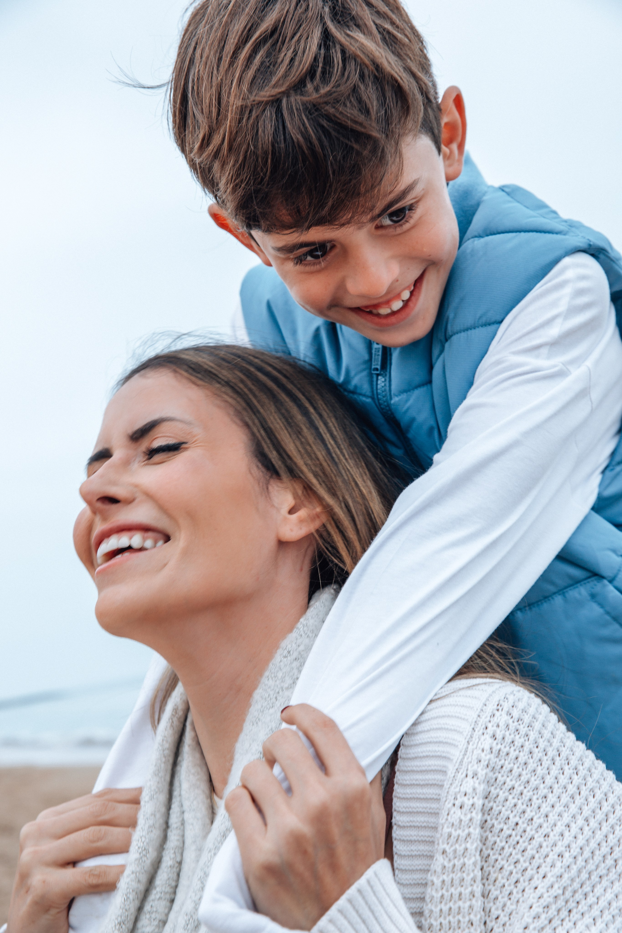 Primer plano de una madre riendo con su hijo abrazándola por detrás — un momento lleno de alegría durante una sesión de fotos familiar en la playa de Denia, España.