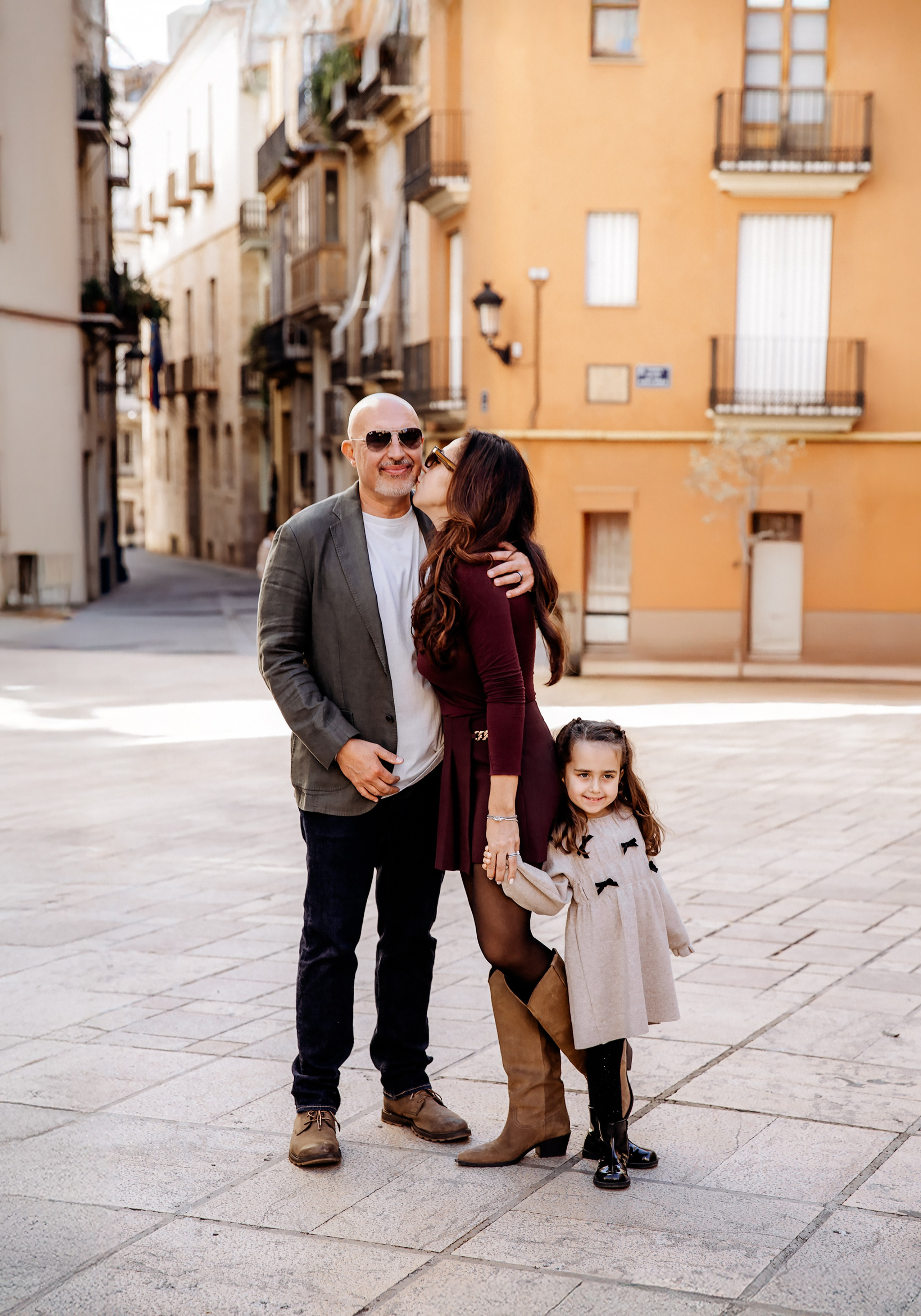 Семейная фотосессия в Валенсии, Испания, где мама, папа и маленькая дочь стоят вместе на уютной площади старого города, снято в тёплом естественном свете на фоне средиземноморской архитектуры — идеально для семейных фотосессий в Валенсии и по всей Испании.