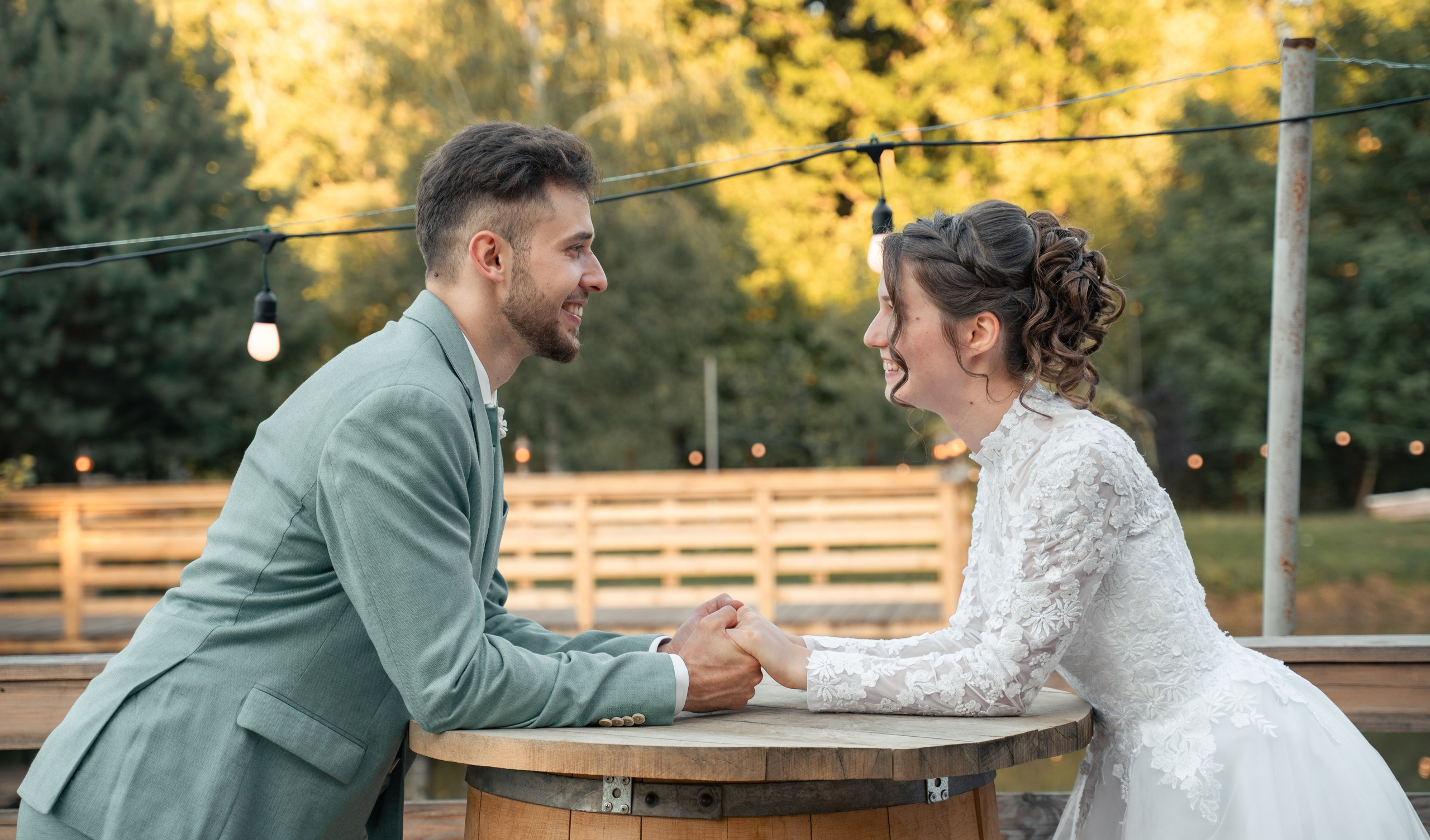 Miri ținându-se de mână la o masă de lemn în aer liber, portret de cuplu la apus pe malul lacului, incadru.ro.