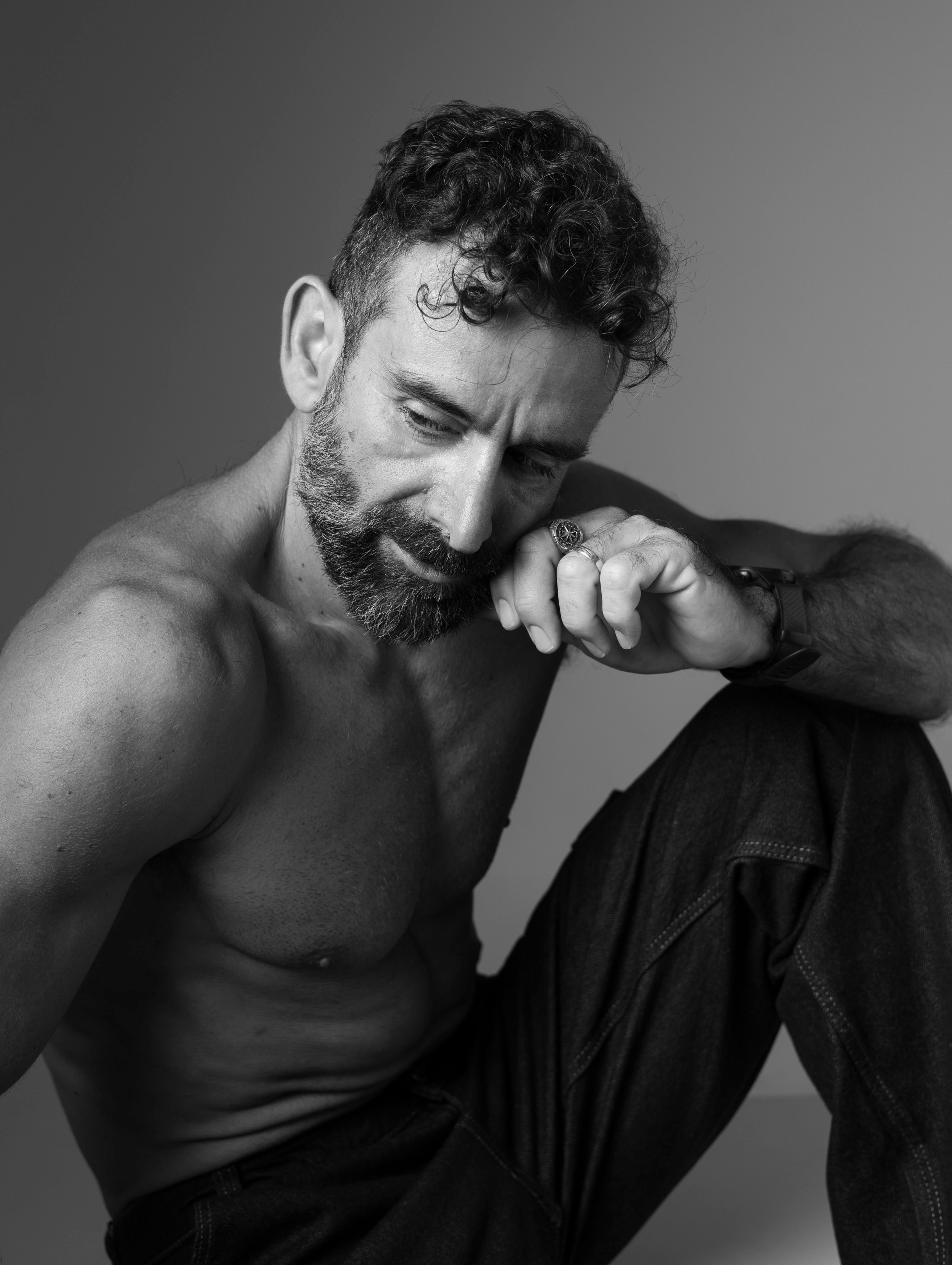 Black-and-white portrait of a shirtless man seated with his hand near his face