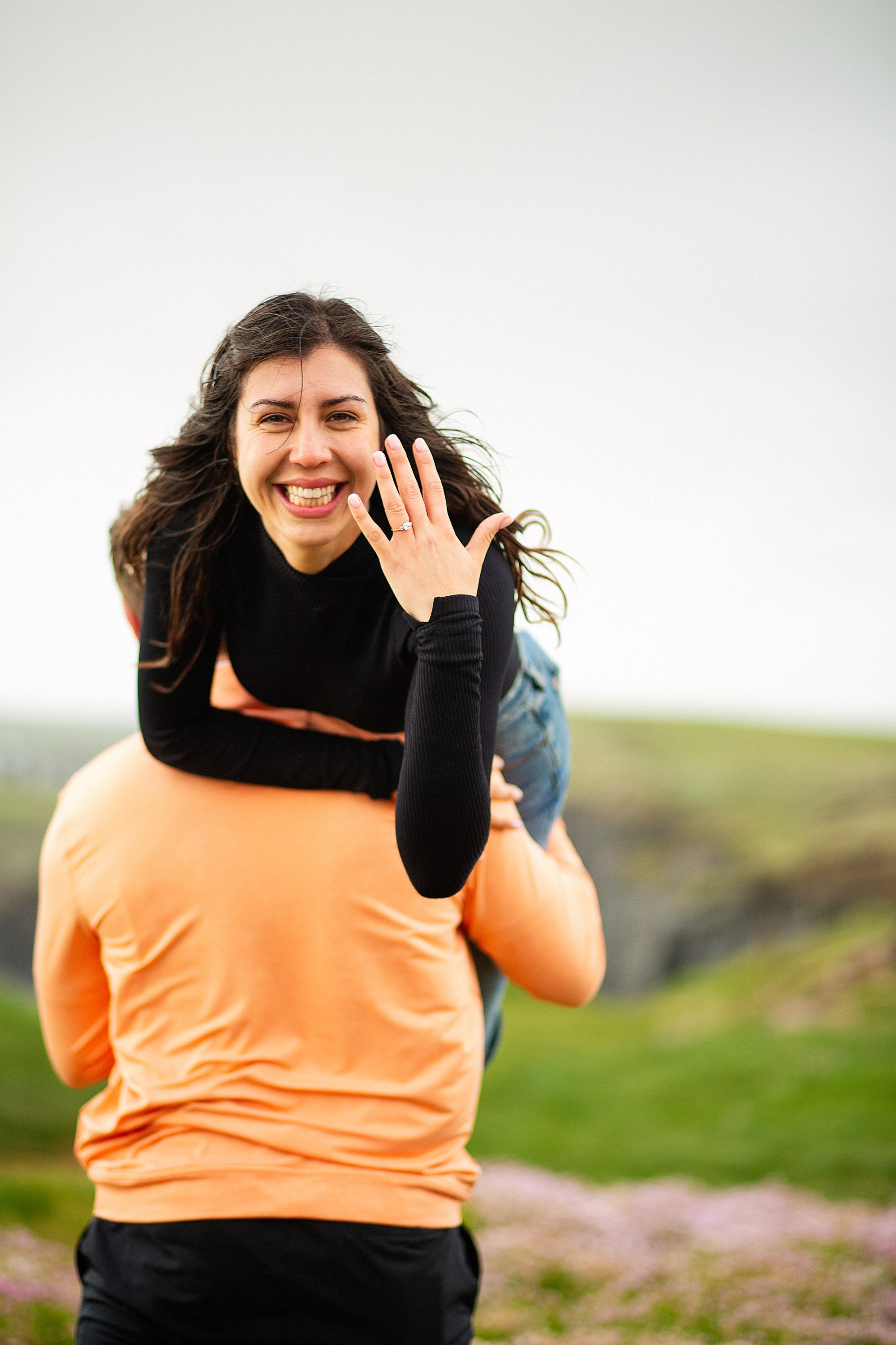 A playful moment of a woman getting a piggyback ride from her partner while showing off her engagement ring in County Galway.