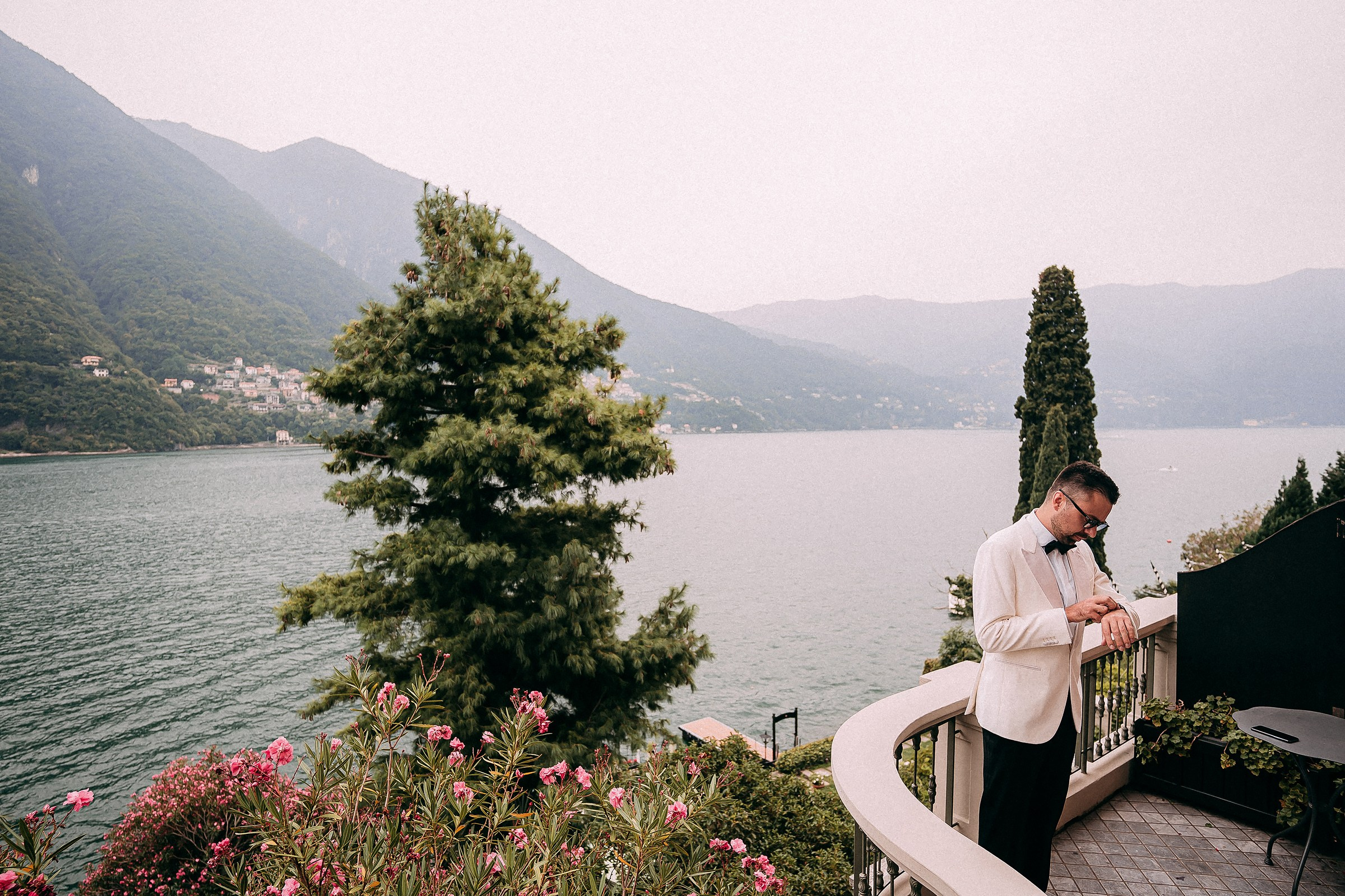 The groom in a white jacket and dark trousers stands on a curved balcony adorned with pink flowers, overlooking a calm lake and lush green mountains under an overcast sky.