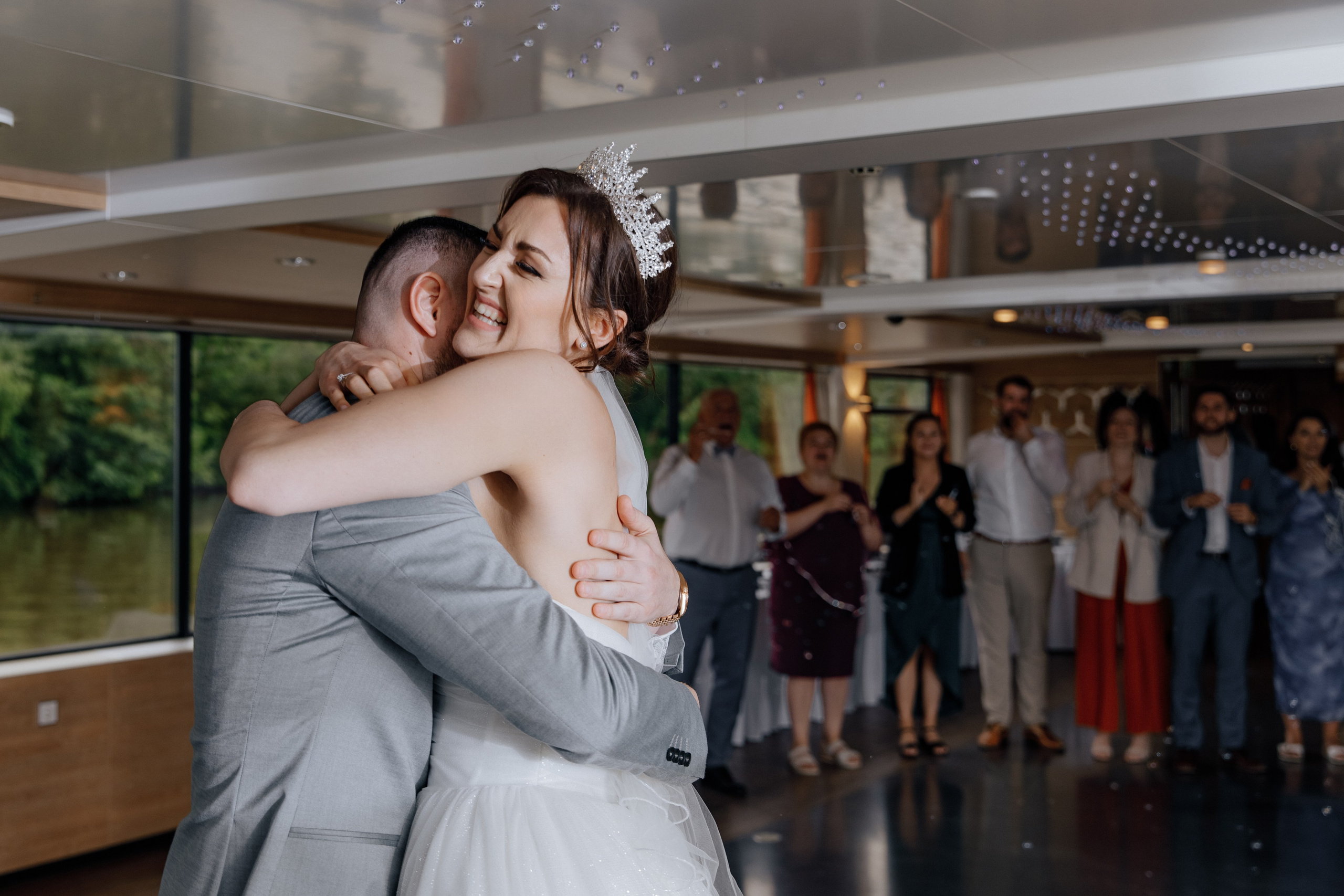 Die Braut umarmt den Bräutigam, während die Gäste im Hintergrund lächeln.