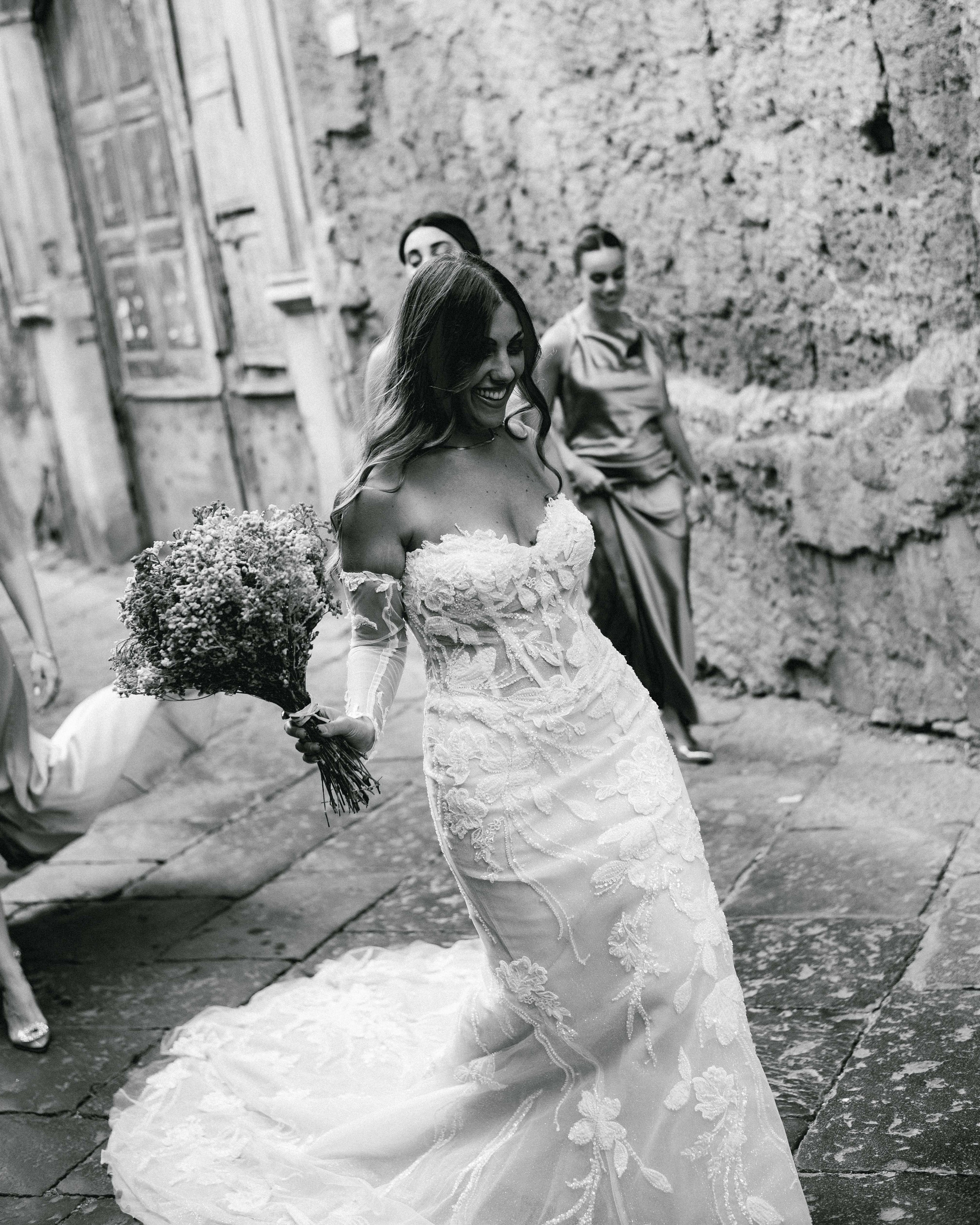 Bride walking through a village holding a bouquet, bridesmaids following behind