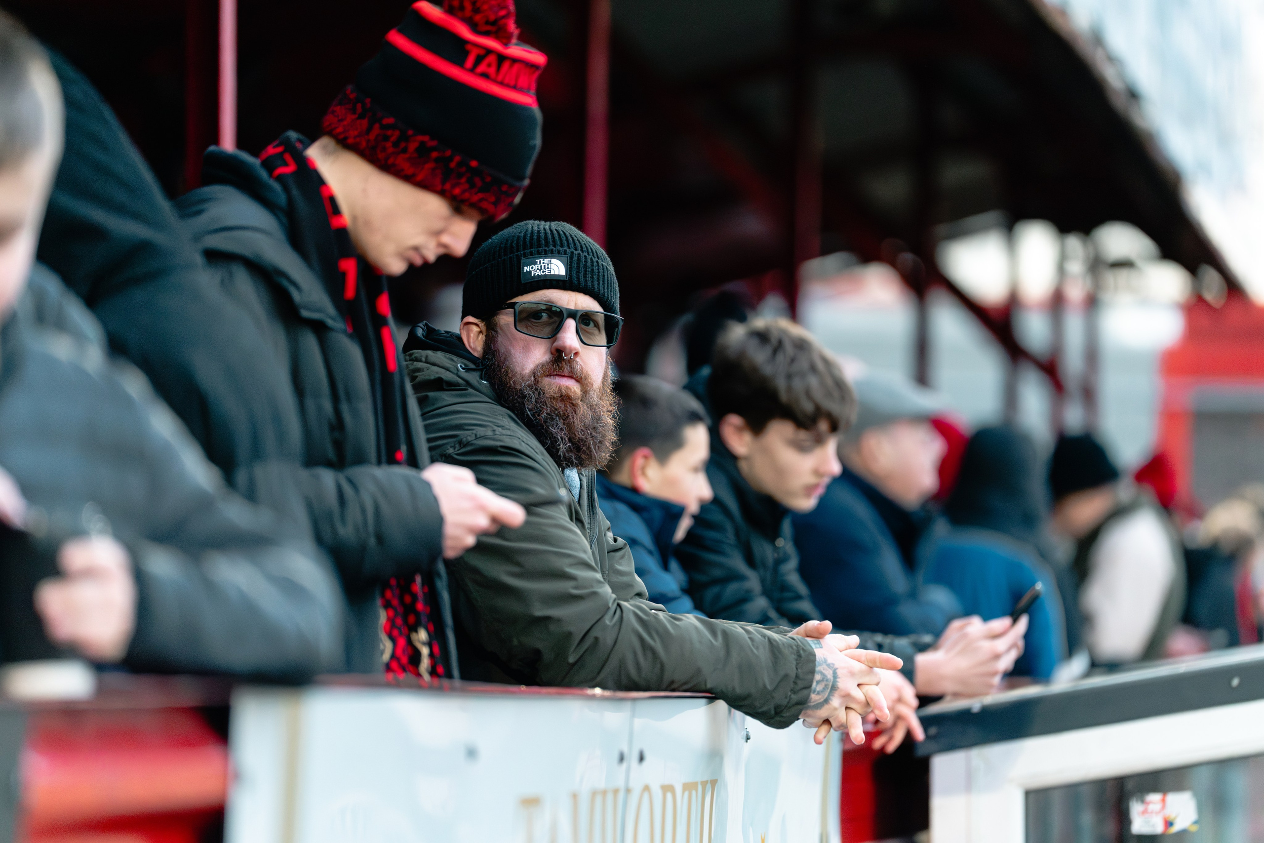 Tamworth supporters in winter clothing lean on the barrier and watch the match from the terrace