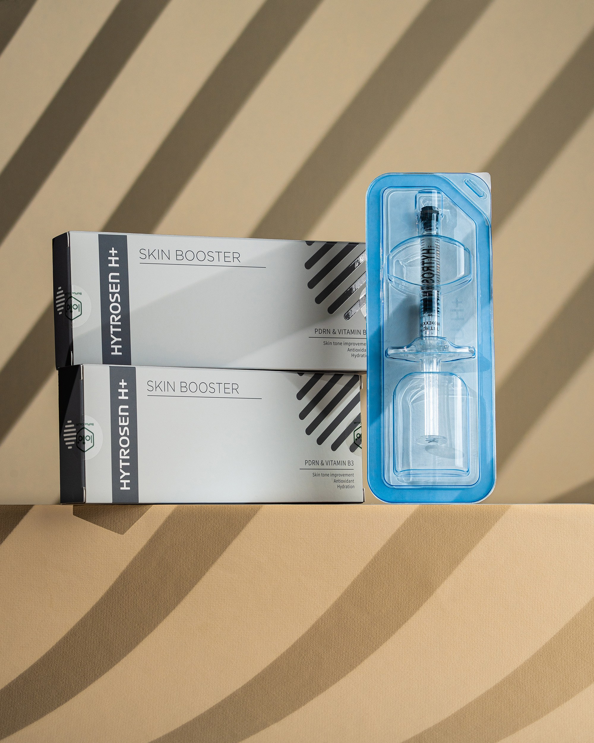 Two grey boxes of HYTROSEN H+ skin booster stacked horizontally with a pre-filled syringe in a blue blister pack standing vertically against a tan shadowed background. Labels highlight PDRN & Vitamin B3 content.