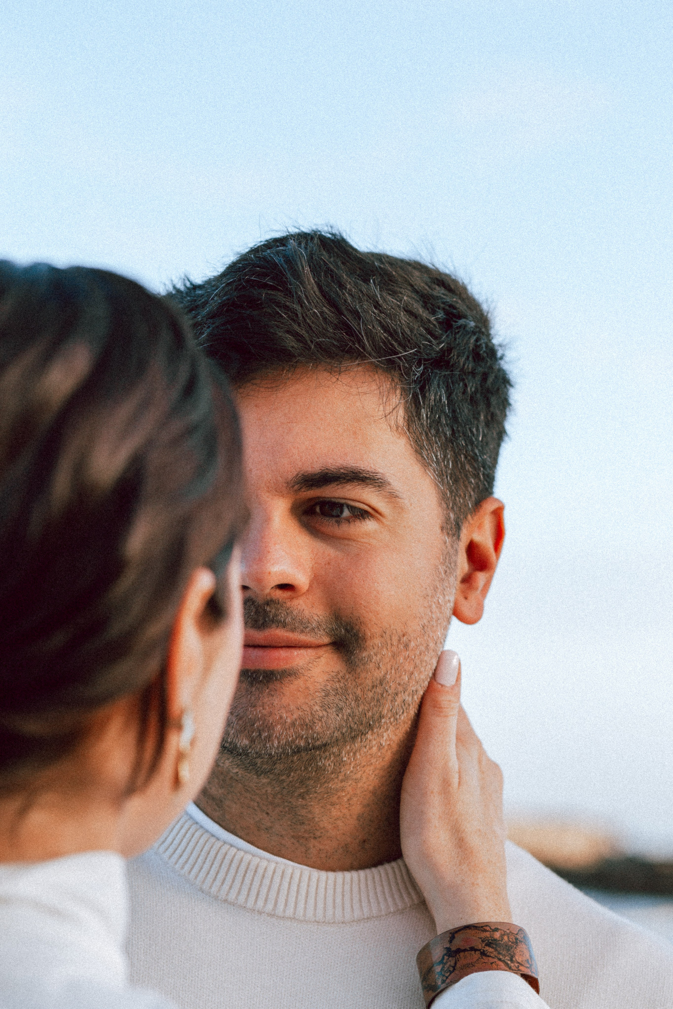 Close-up portrait of a couple sharing an intimate gaze, with the man smiling softly as his partner gently touches his face, set against the soft blue sky during golden hour in Valencia, Spain.