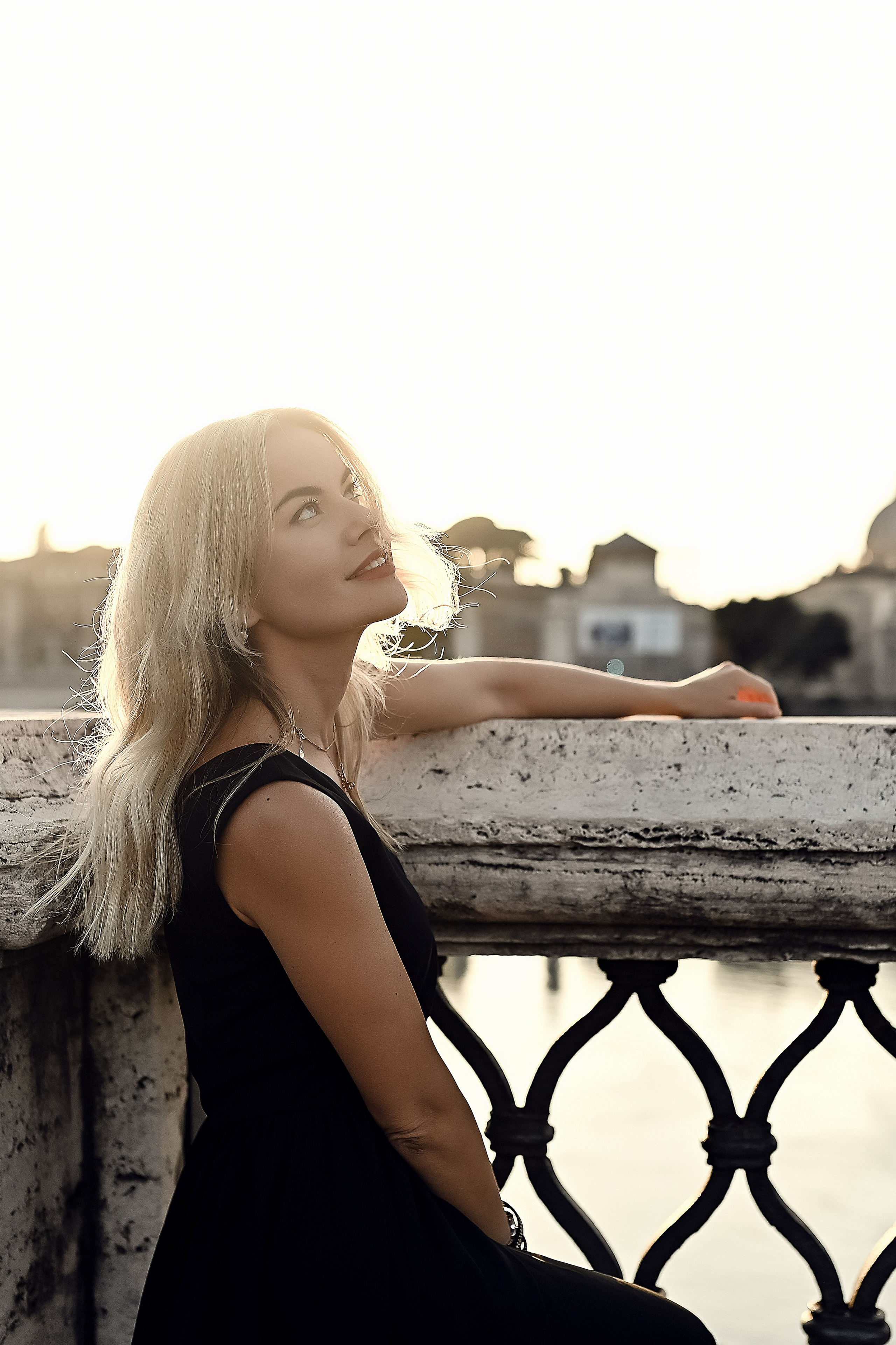 A blonde woman in a black dress leans against a stone balustrade at sunset, gazing dreamily at the sky.