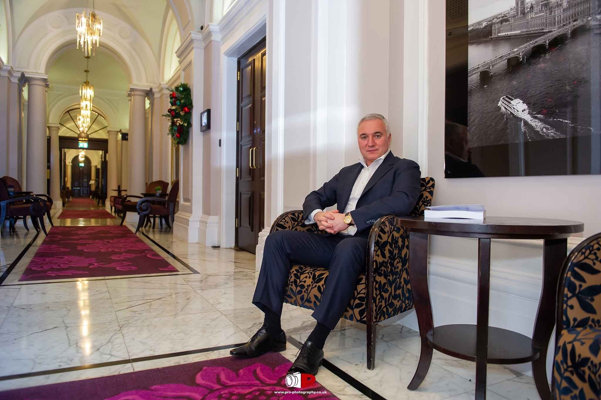 The businessman is seated in a luxurious hotel hallway with elegant furnishings and marble floors.