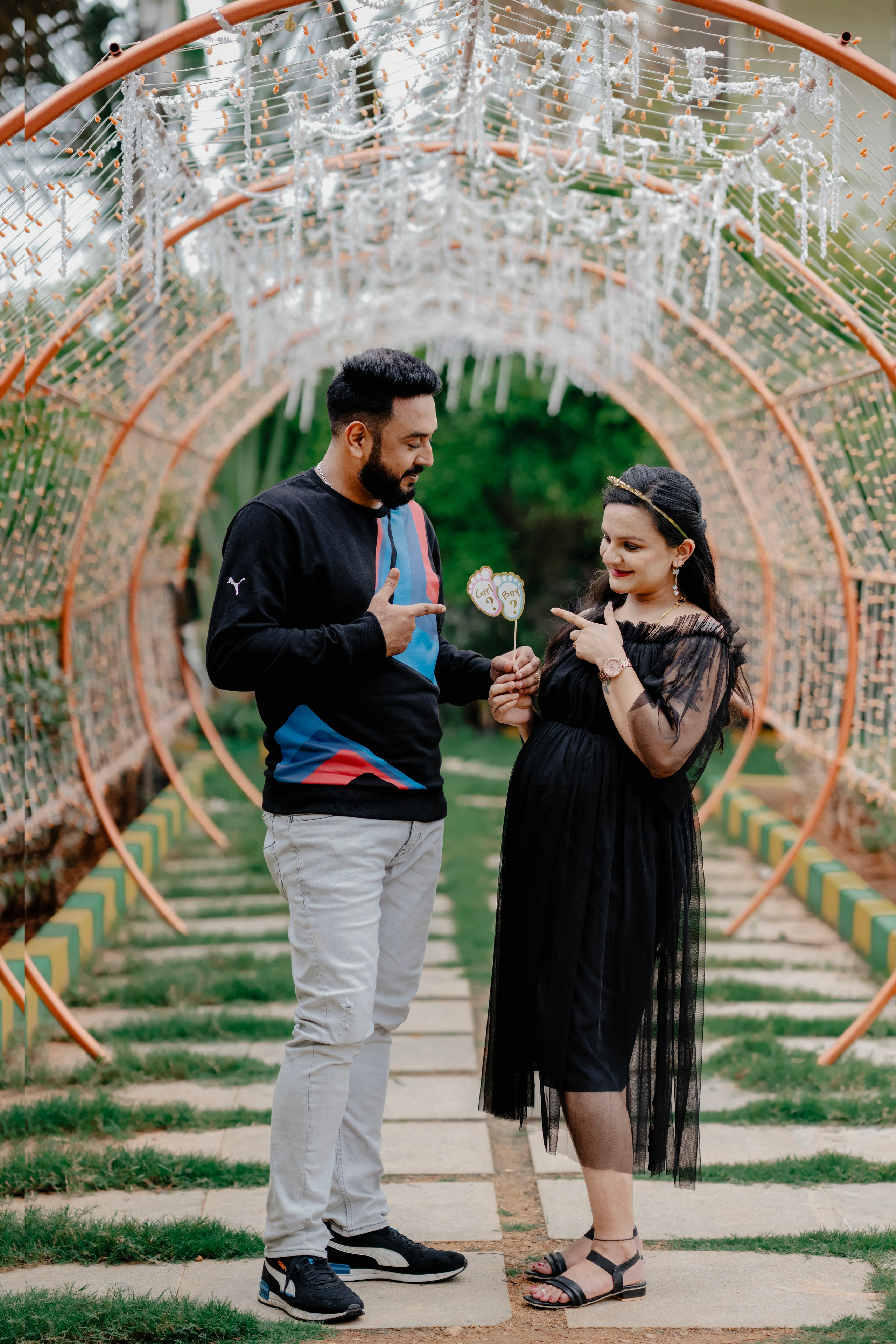 Outdoor maternity photoshoot in Bengaluru featuring a couple in black outfits pointing at baby feet props inside a decorated garden archway tunnel.