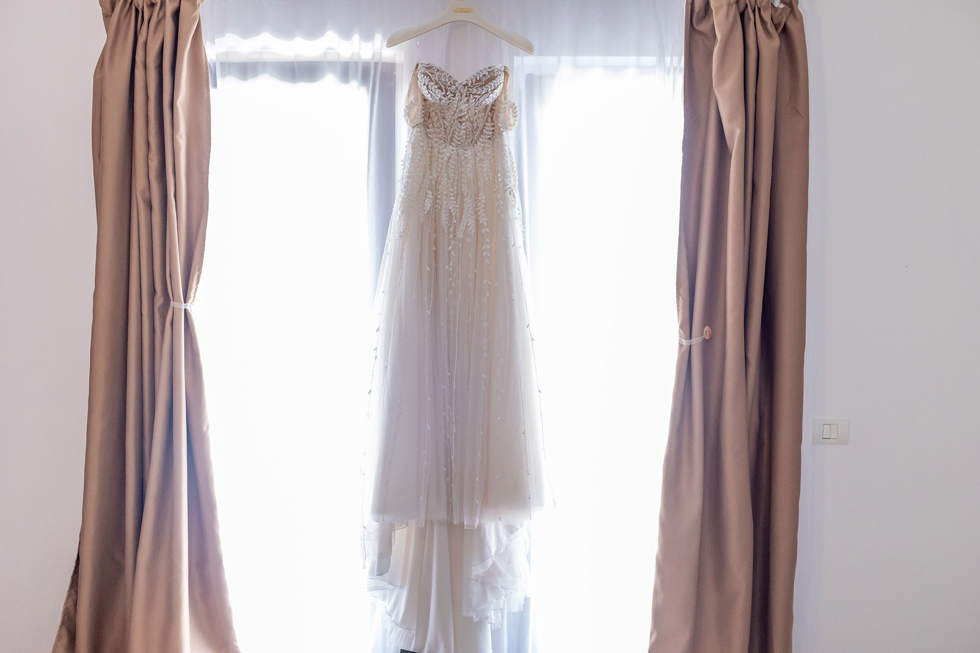 Rochie de mireasă fină cu broderie florală și dantelă, agățată pe umeraș în fața unei ferestre luminoase încadrate de perdele bej.