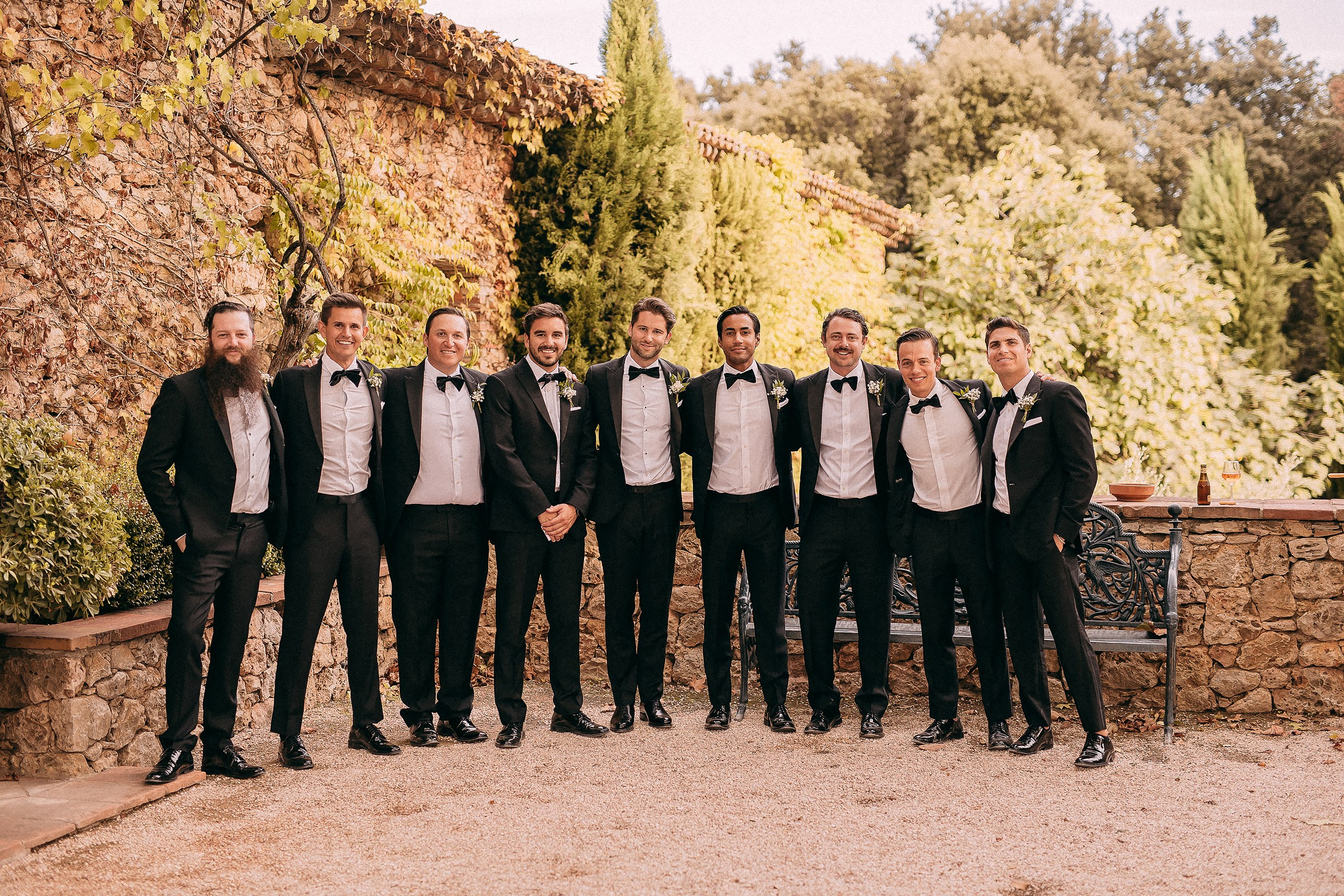 A formal yet candid photo of the groom and his groomsmen standing in a row, all dressed in sharp black tuxedos, against a rustic stone backdrop.