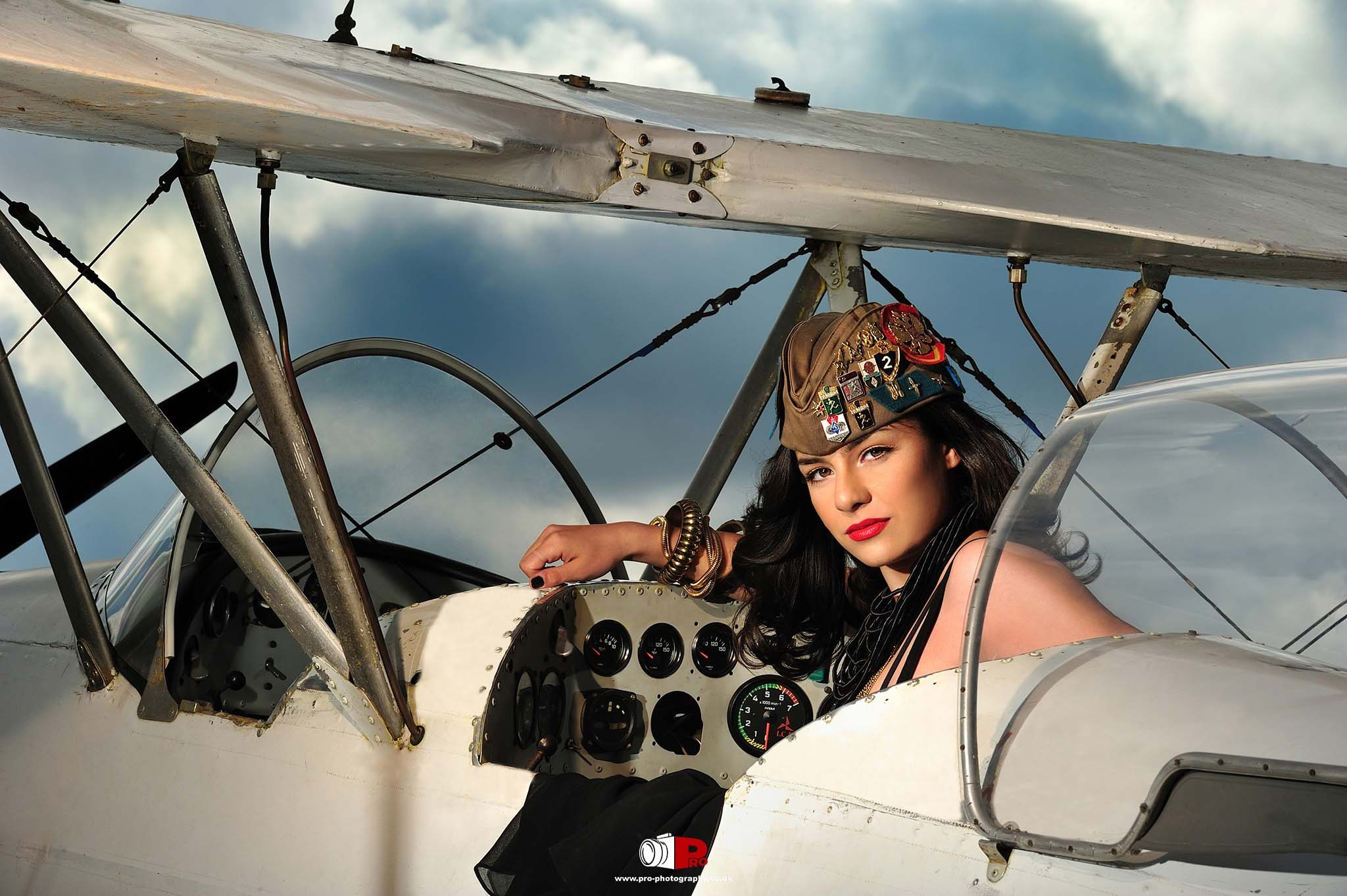 A woman with red lips and a vintage military cap posing inside an old aeroplane cockpit against a dramatic sky.