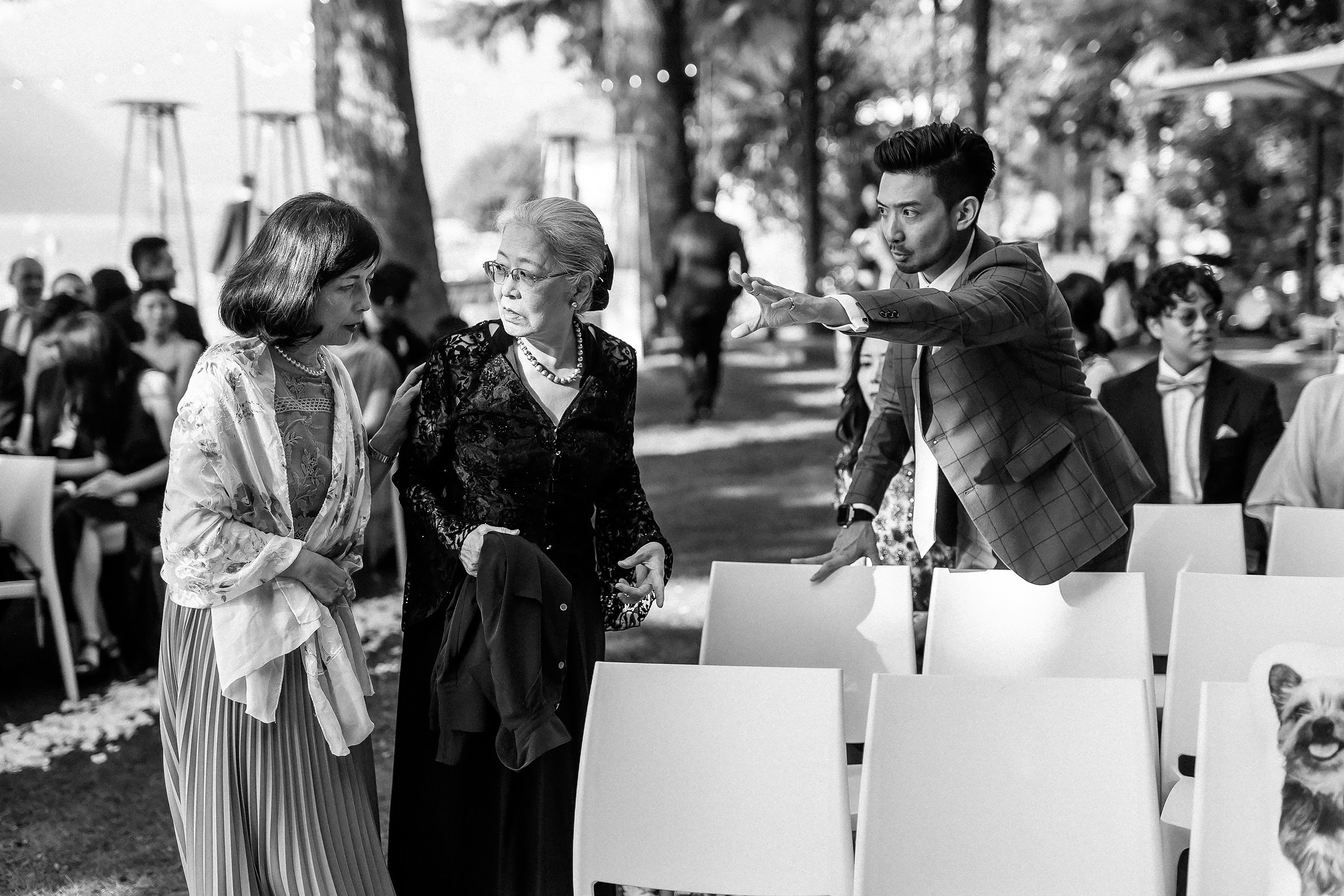Wedding guest in plaid jacket gestures to two elderly women, helping them find their seats.