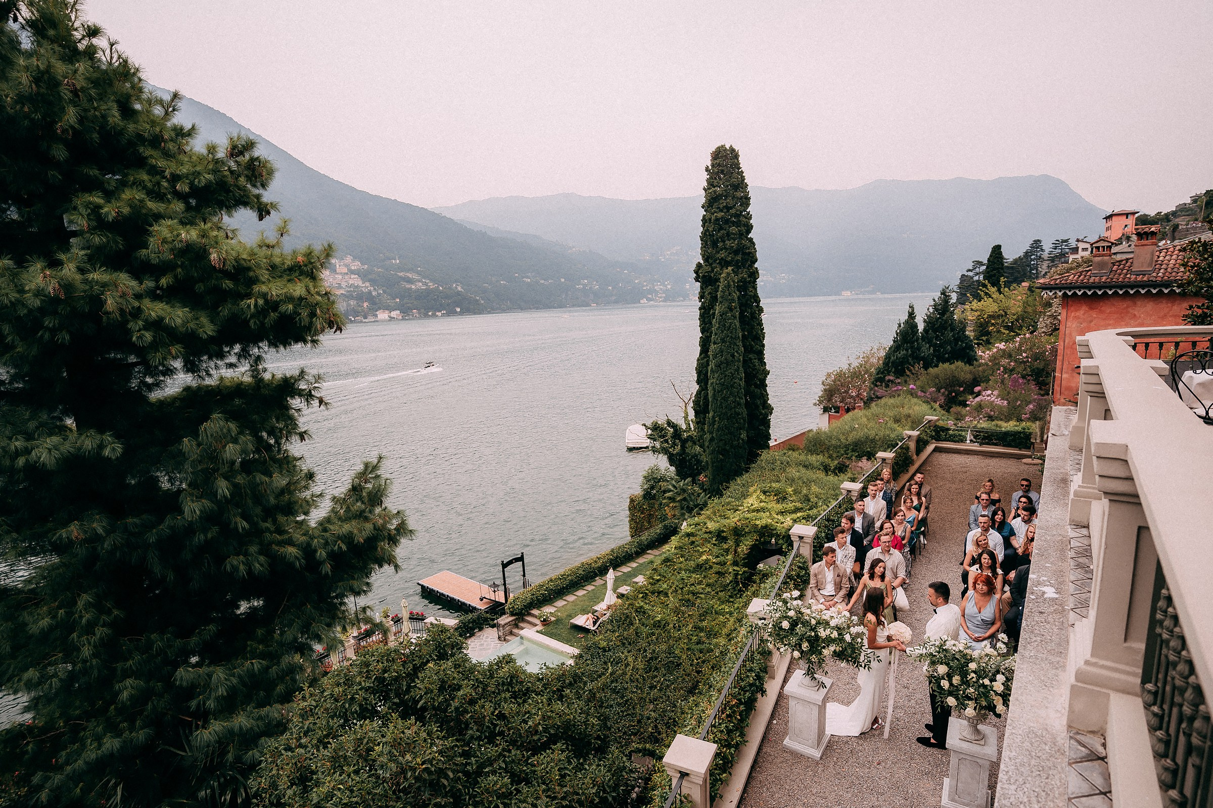 Romantic lakeside wedding ceremony with mountain views of Relais Villa Vittoria, floral altar, and seated guests in a serene outdoor setting. 