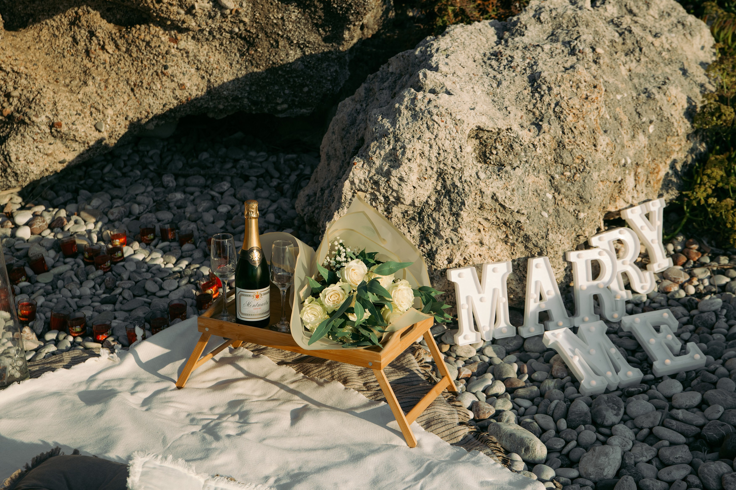"Der Schriftzug 'MARRY ME' am Strand von Kato Petres während der Golden Hour. Die weißen Buchstaben leuchten vor dem tiefblauen Ägäischen Meer, während die Brandung sanft gegen die Steine schlägt. Das warme Licht des Sonnenuntergangs spiegelt sich in den Wellen und auf den Felsen