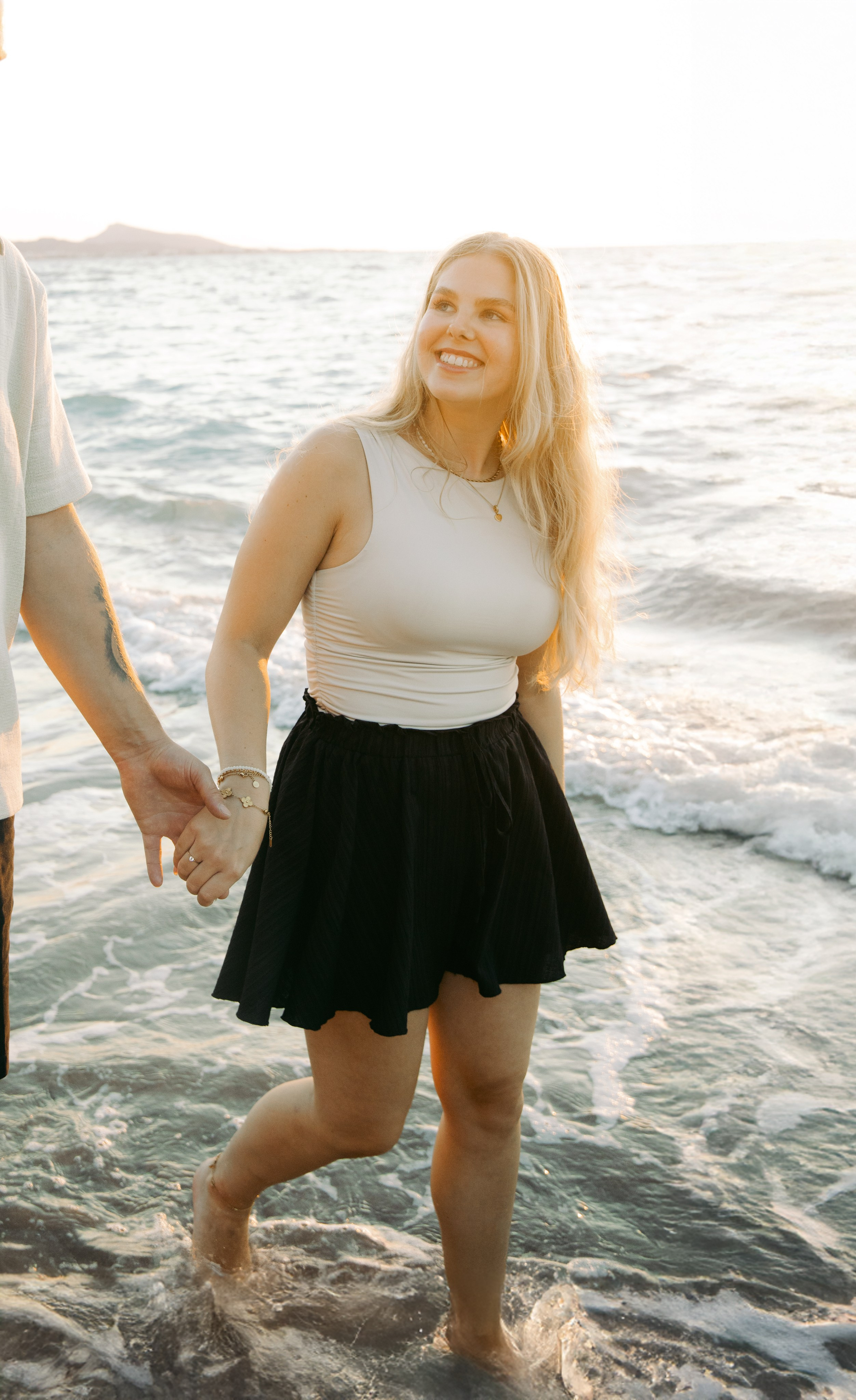 "Hand in Hand am Kato Petres: Detailaufnahme zweier sich haltender Hände über dem steinigen Ufer. Ein Symbol der Zweisamkeit vor der wilden, natürlichen Schönheit der Westküste von Rhodos."