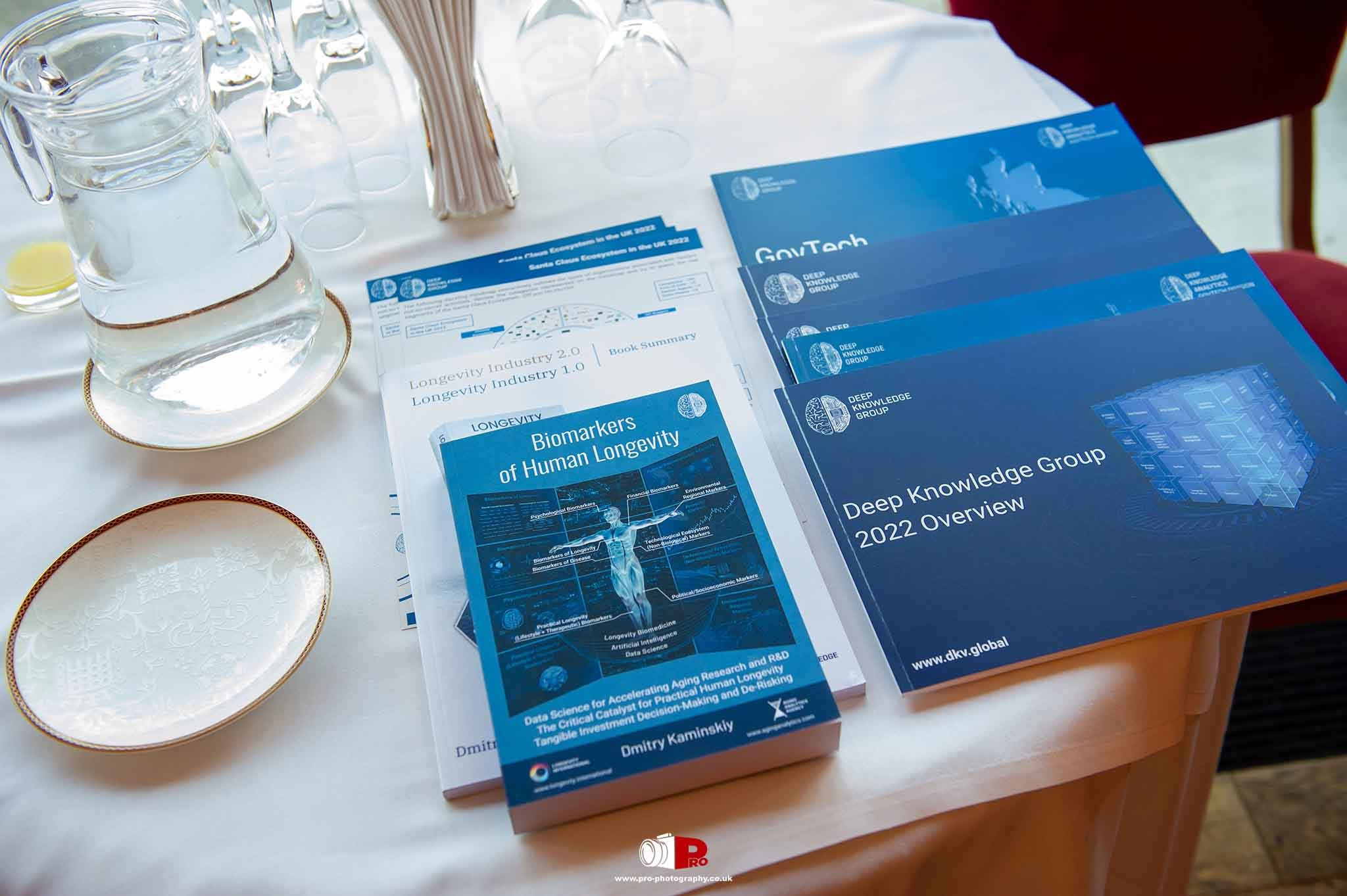 Table display with informational booklets, brochures, and resources for a Deep Knowledge Group business event.