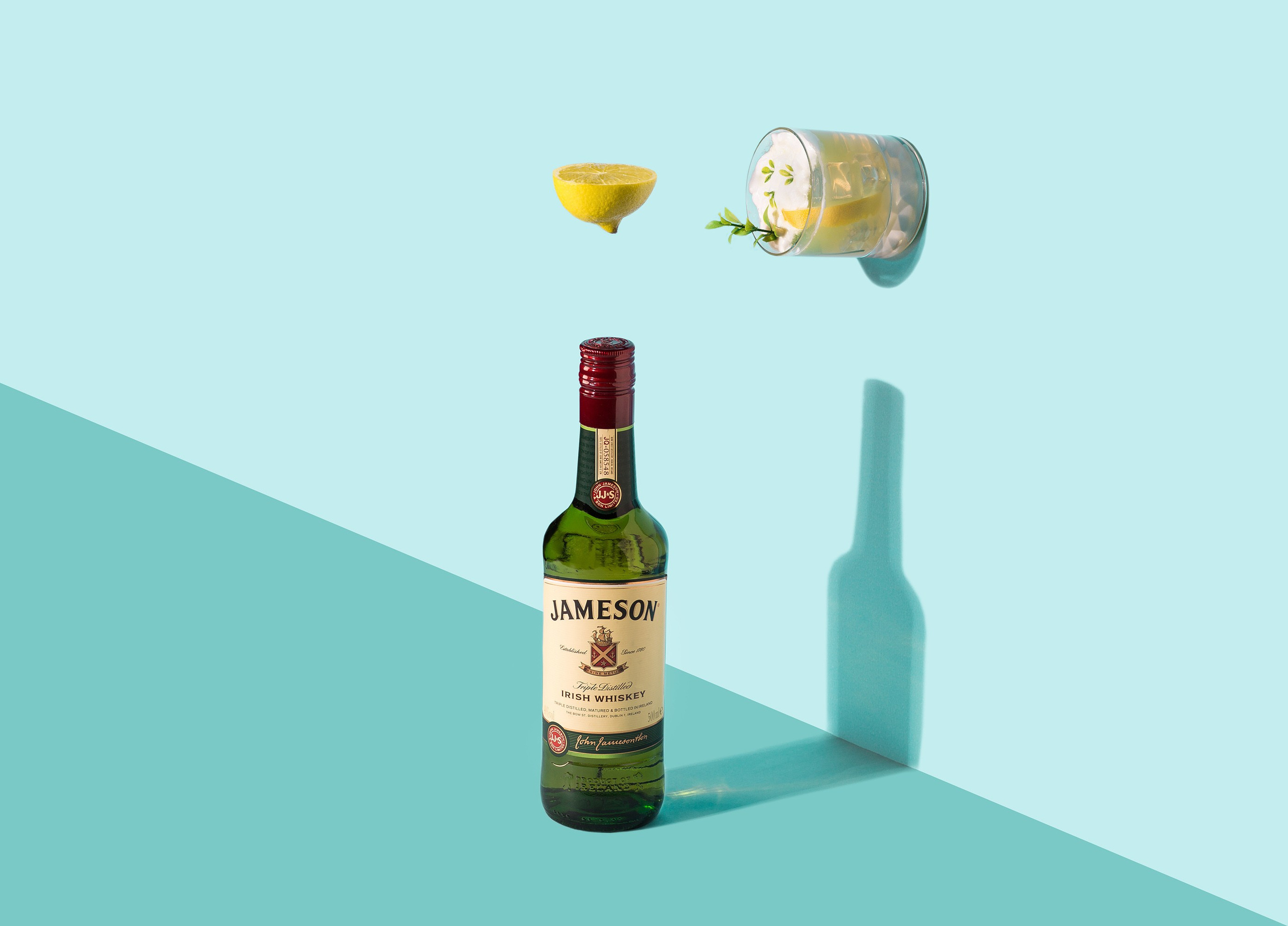 Jameson viskisi şüşəsi, yuxarıda isə limon dilimi və içki olan kokteyl stəkanı yerləşdirilmiş minimalistik, mavi fonda çəkiliş.