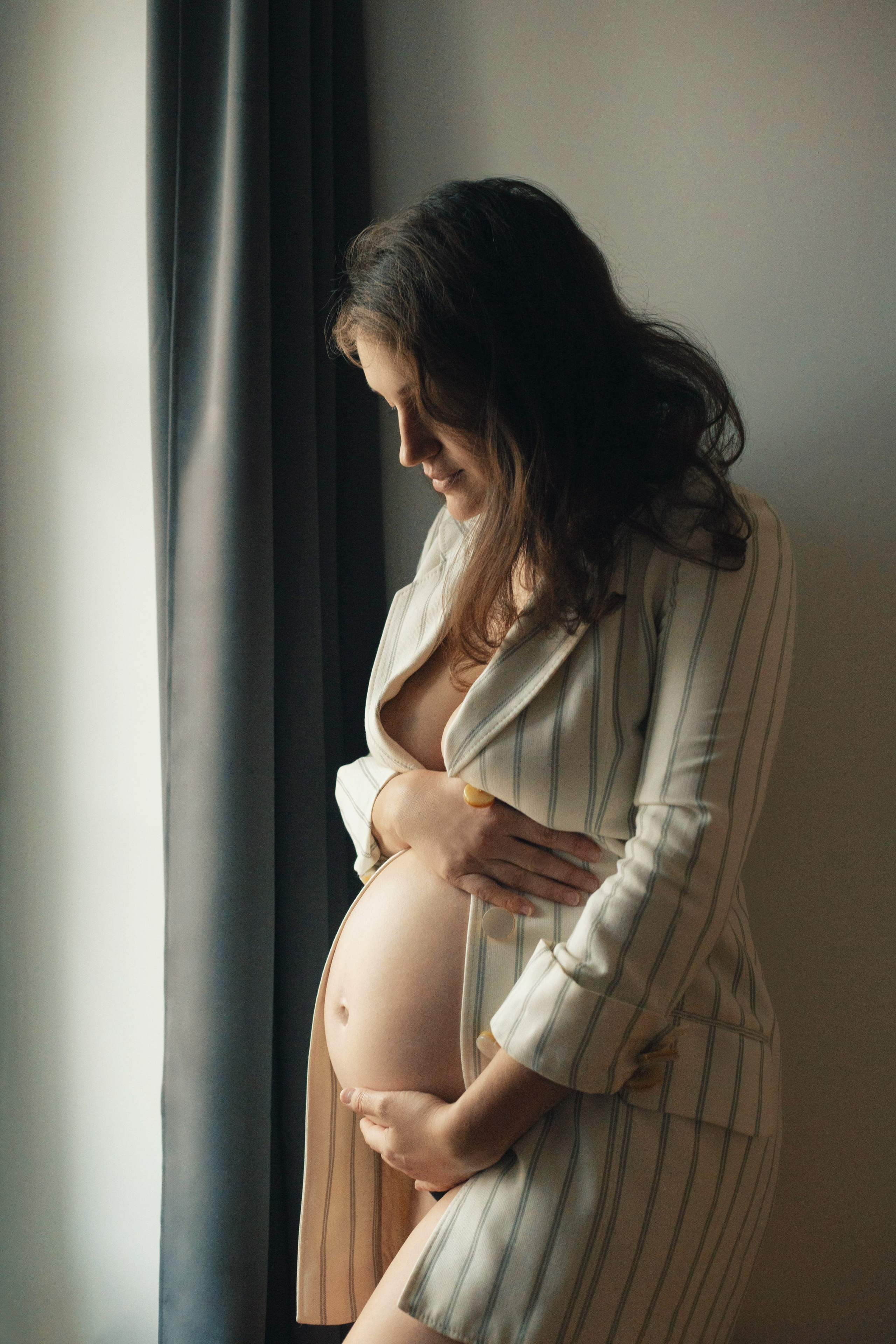 Femme enceinte douce en robe beige regardant tendrement son ventre près de la fenêtre, lumière naturelle