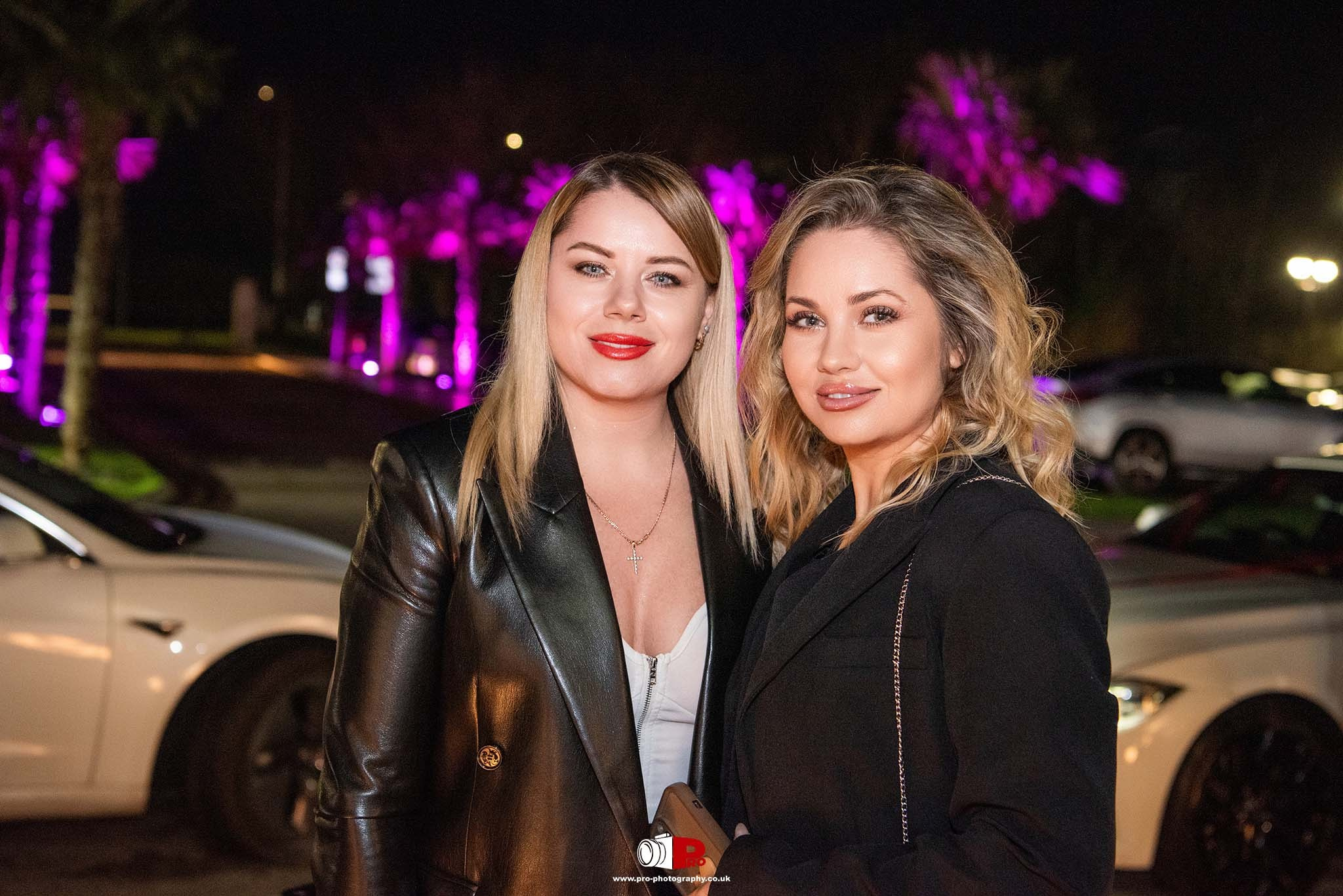 Two women in stylish black outfits are posing together outdoors, with pink lighting and luxury cars in the background.