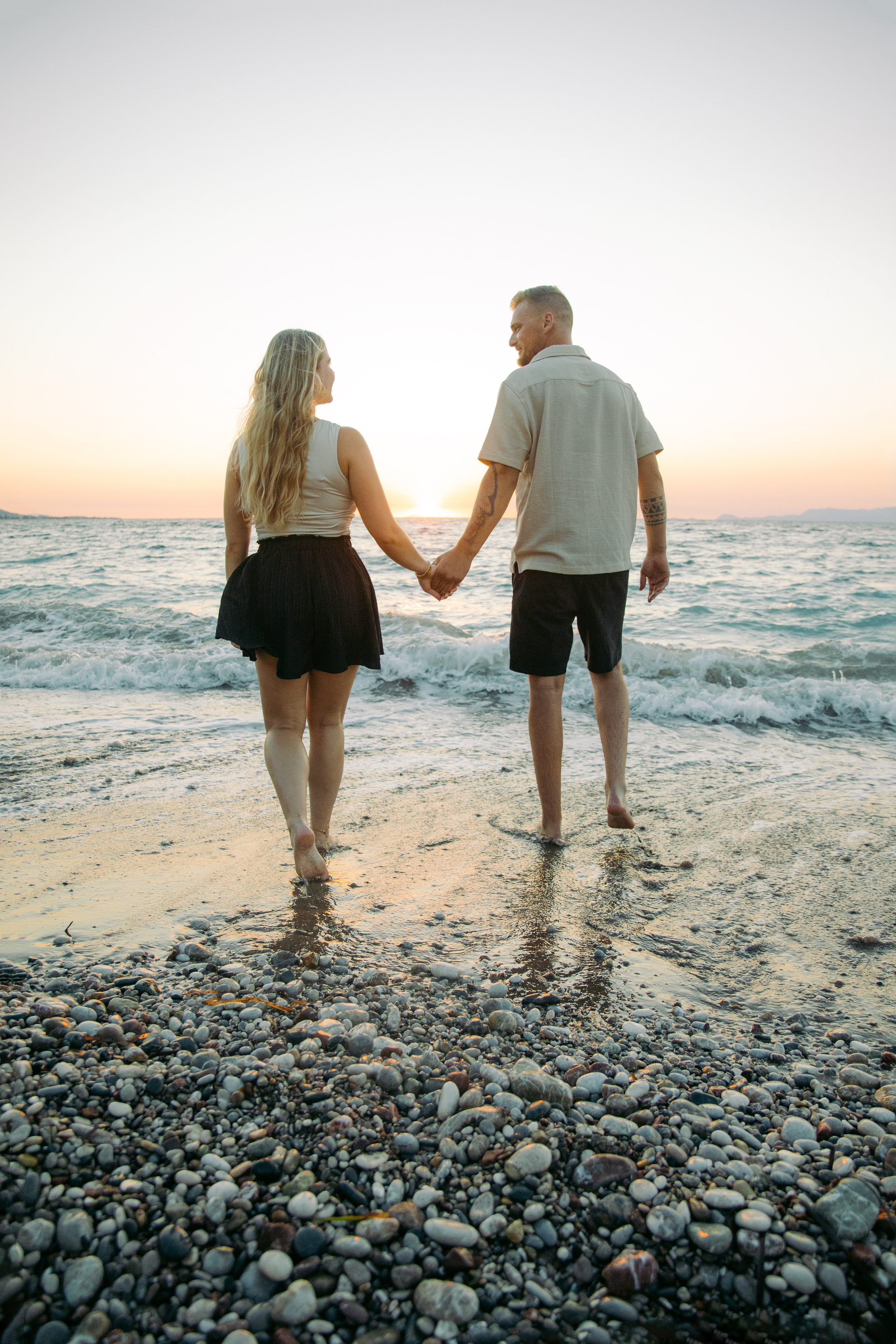 "Ein Paar spaziert Hand in Hand am steinigen Ufer von Kato Petres auf Rhodos entlang. Sie gehen direkt am Wasser, während im Hintergrund die hohen, zerklüfteten Kalksteinfelsen aufragen und das blaue Meer sanft gegen die Kiesel brandet."