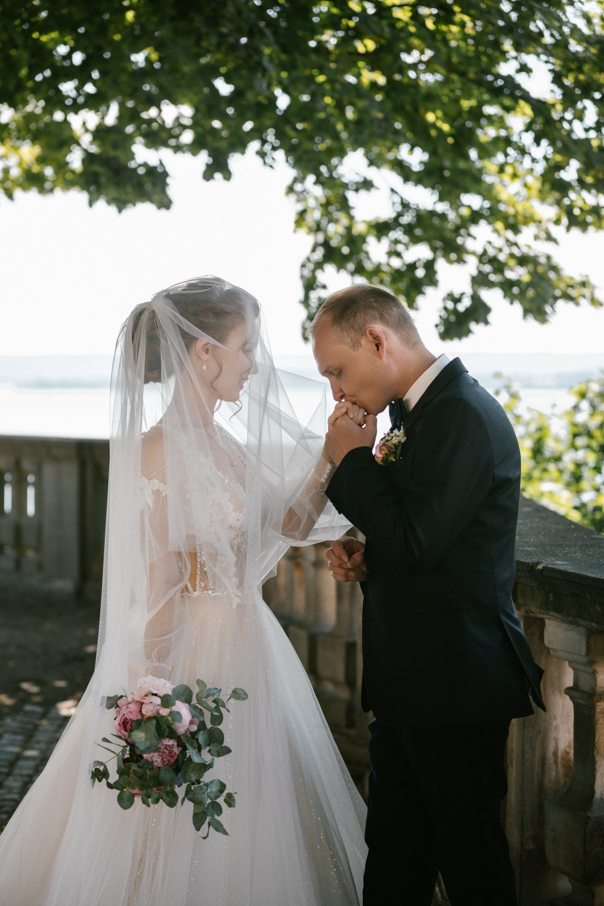 Groom kisses bride’s hand on lakeview balcony at Schloss Meersburg, bouquet visibl
