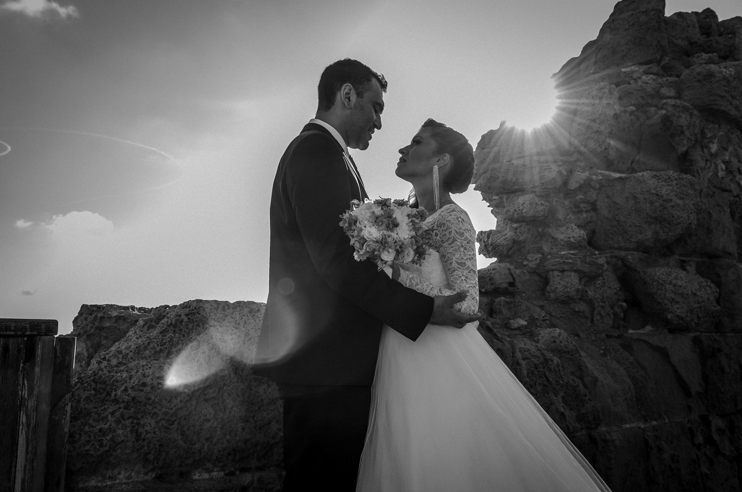 Black and white wedding photography of a bride and groom embracing against ancient stone ruins with sun flare in Caesarea, Israel