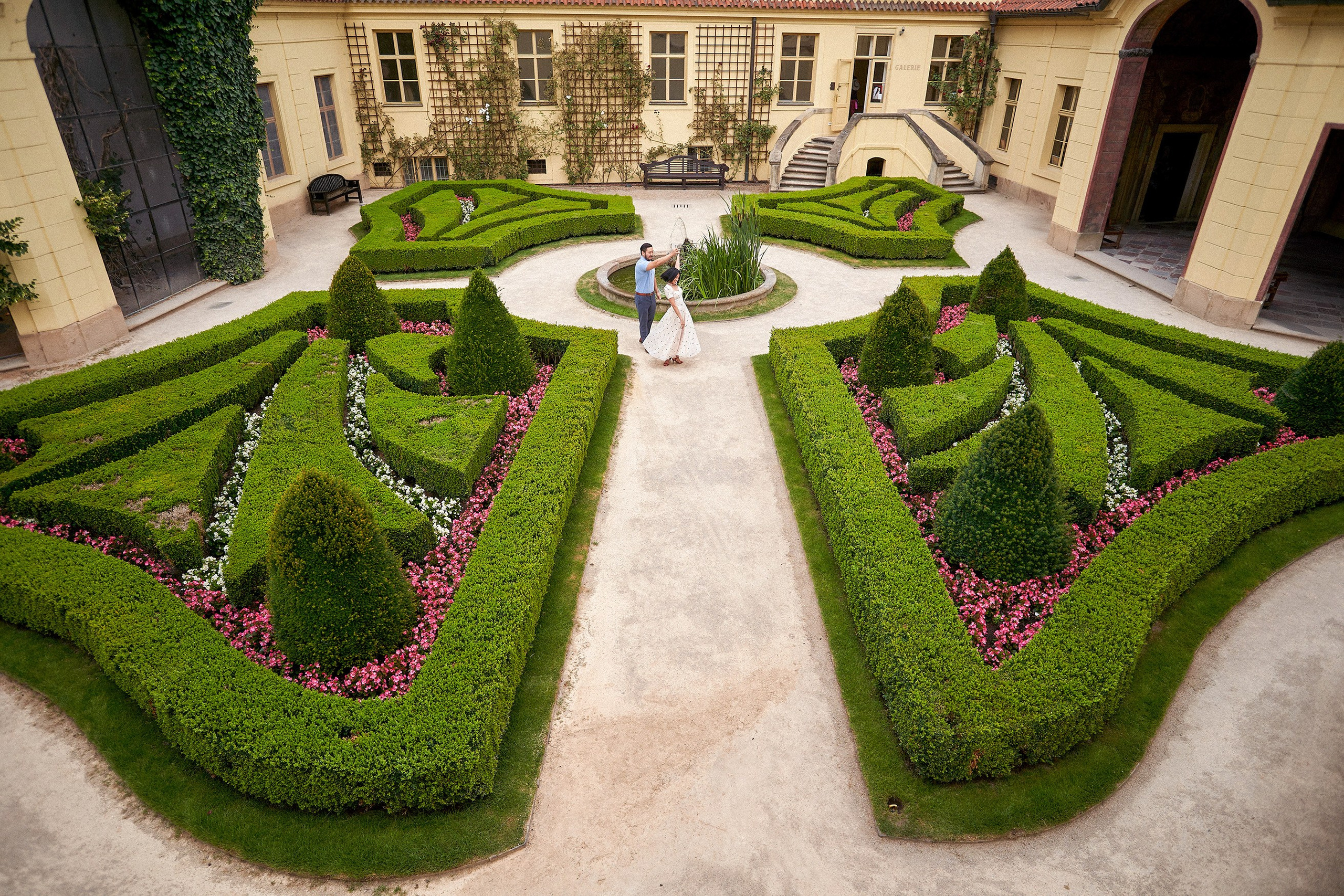 Aerial view of engaged couple walking hand-in-hand through the manicured grounds of Vrtba Garden, Prague.
