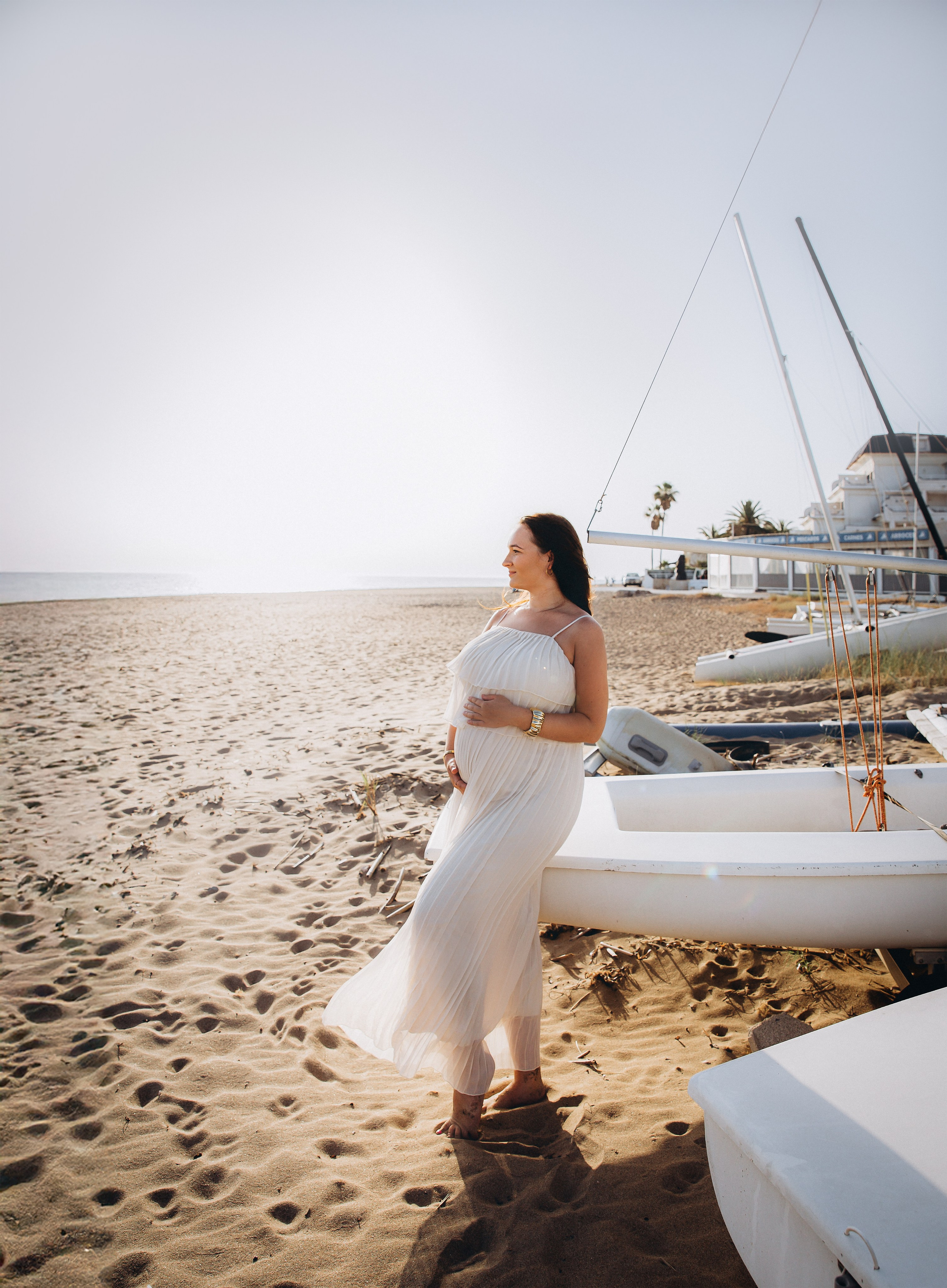 Mujer embarazada con vestido blanco fluido posando junto a veleros en la playa de Denia, España — sesión de maternidad serena y elegante para quienes buscan fotografía natural junto al mar en la Costa Blanca.