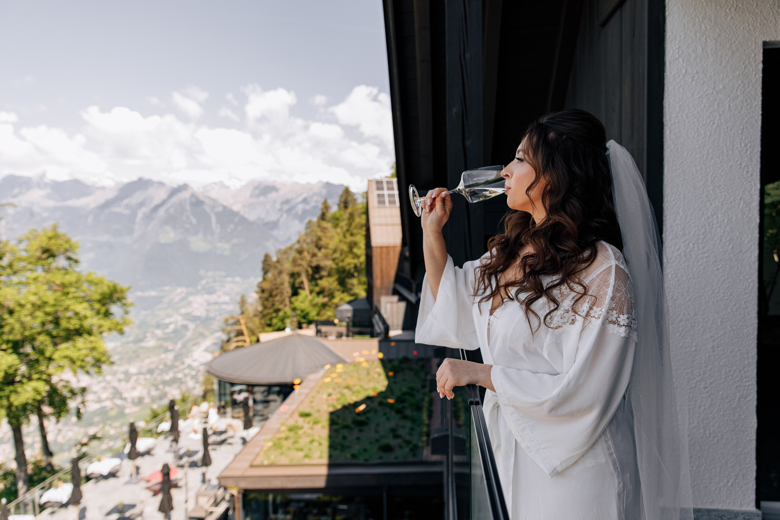 Die Braut steht auf dem Balkon eines Hotels und trinkt ein Glas Wein mit Blick auf die Tiroler Berge. 