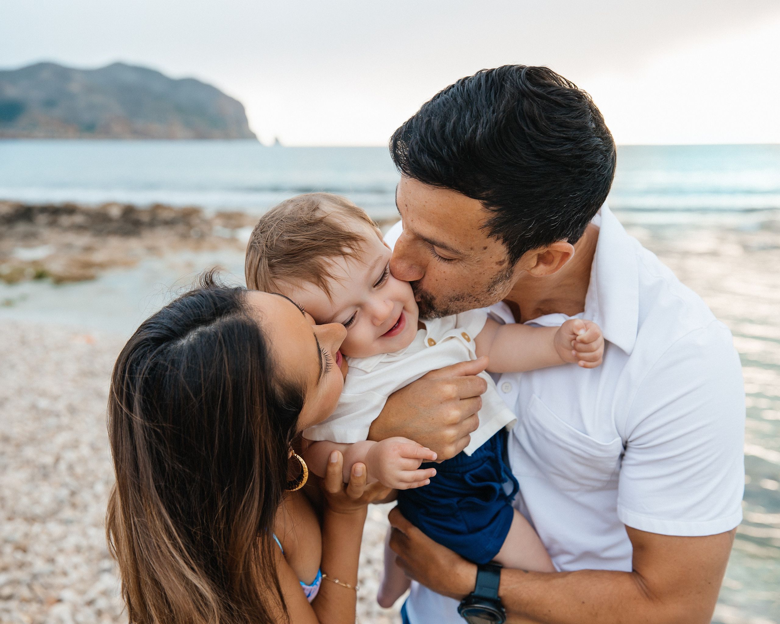 Sesión de fotos familiar en Alicante, España, con unos padres cariñosos besando a su pequeño hijo en una hermosa playa del Mediterráneo con espectaculares vistas al mar. Un emotivo retrato familiar lifestyle que transmite amor, felicidad y recuerdos inolvidables de sus vacaciones. Ideal para fotografía familiar, sesiones familiares en la playa, retratos infantiles y fotografía familiar de destino en Alicante y toda España.