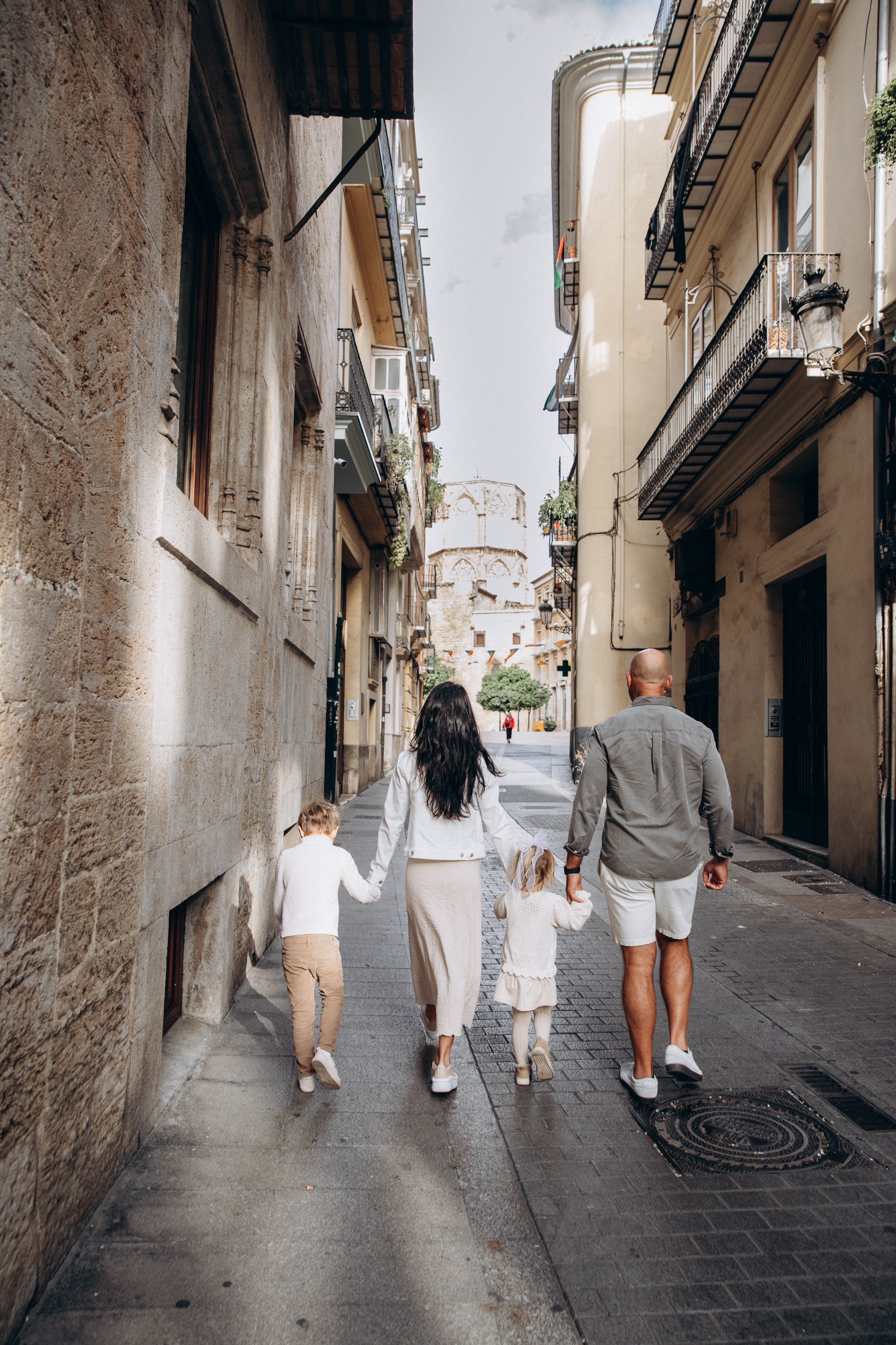 Sesión de fotos familiar en Valencia, España, con una familia de cuatro caminando de la mano por una encantadora calle histórica del casco antiguo de Valencia, rodeada de la clásica arquitectura mediterránea. Un retrato familiar lifestyle espontáneo que captura unión, amor y recuerdos inolvidables de sus vacaciones. Ideal para fotografía familiar, sesiones de fotos familiares, retratos infantiles, fotografía de viajes y fotografía familiar de destino en Valencia y toda España.