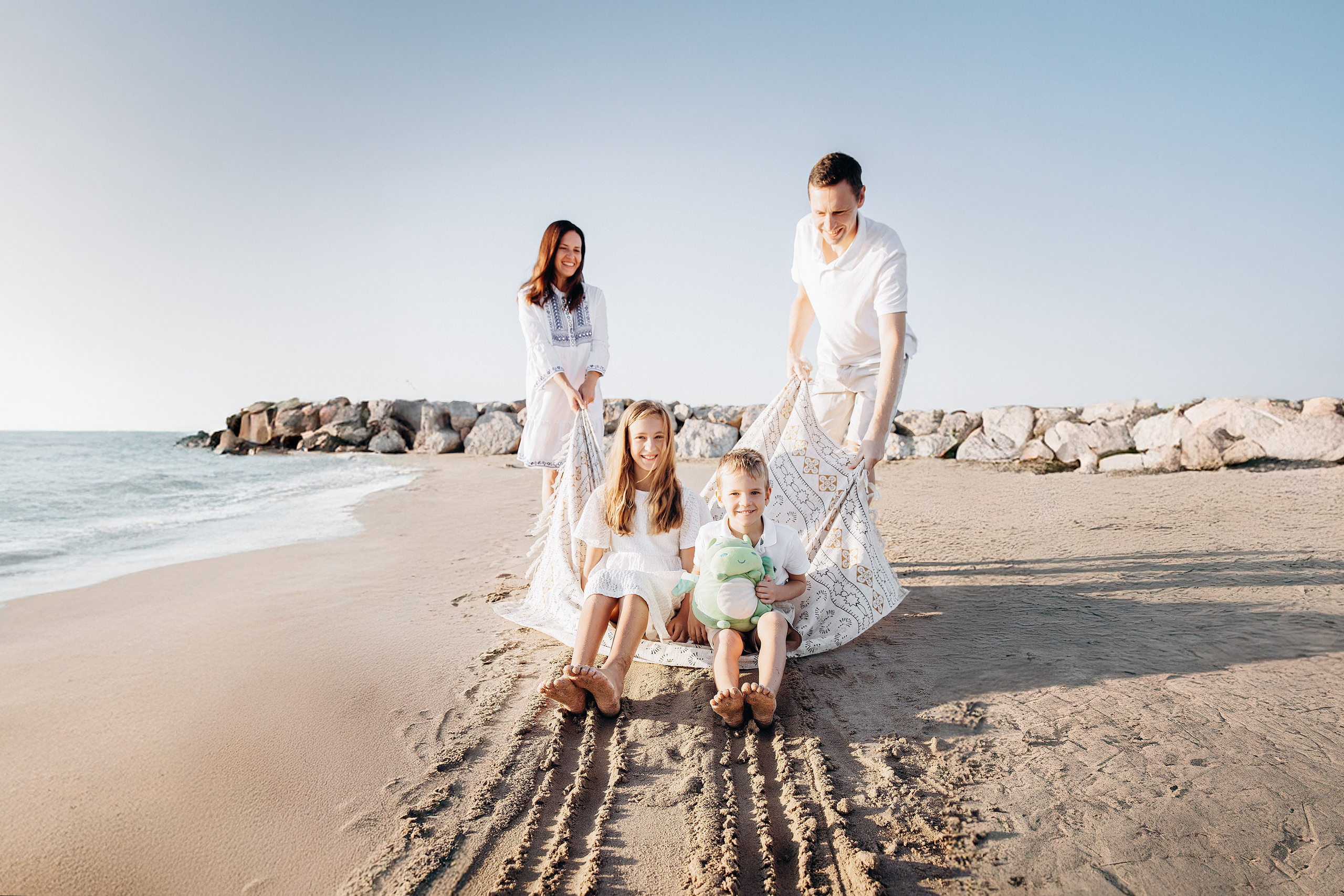 Alegre sesión familiar en la playa de Valencia, España, con una madre tirando de sus hijos sobre una manta en la arena mientras otro familiar los observa — perfecta como inspiración para sesiones familiares relajadas y divertidas en Valencia y la costa española.