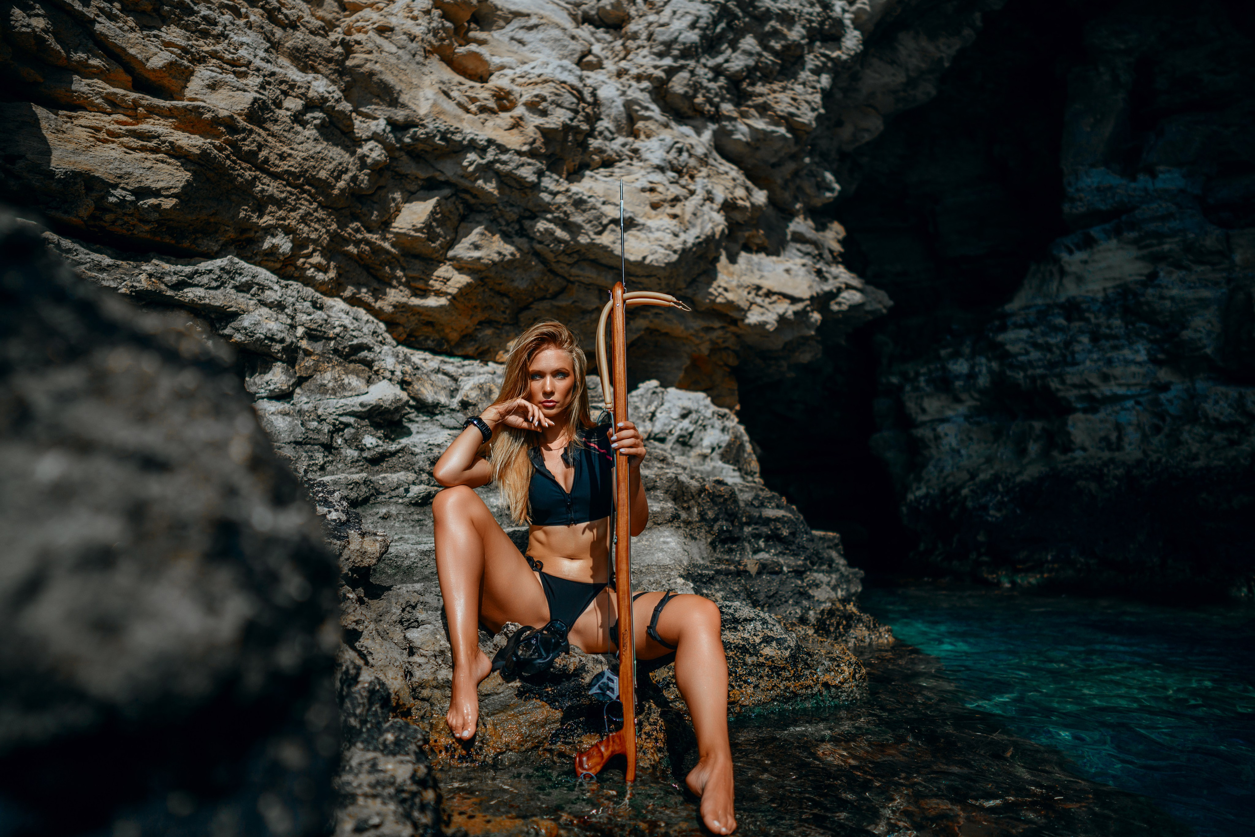 At the ocean's edge, framed by the majestic, towering cliffs, a professional and stylish female photoshoot unfolds. A young woman, adorned in a lithe diving bikini, a sleek underwater hunting rifle cradled in her arms, sits gracefully on the weathered rocks. Her eyes, a window to her adventurous spirit, lock onto the camera, beckoning with intrigue