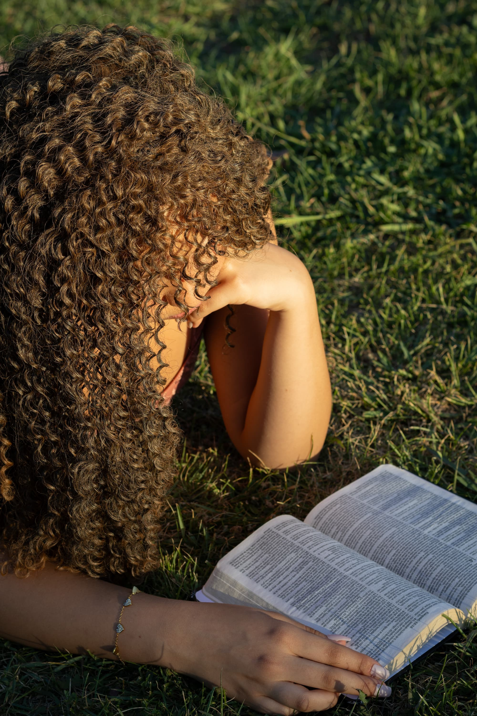 Ensaio fotográfico feminino ao ar livre. Jovem de cabelos cacheados deitada de bruços na grama verde, lendo um livro aberto.