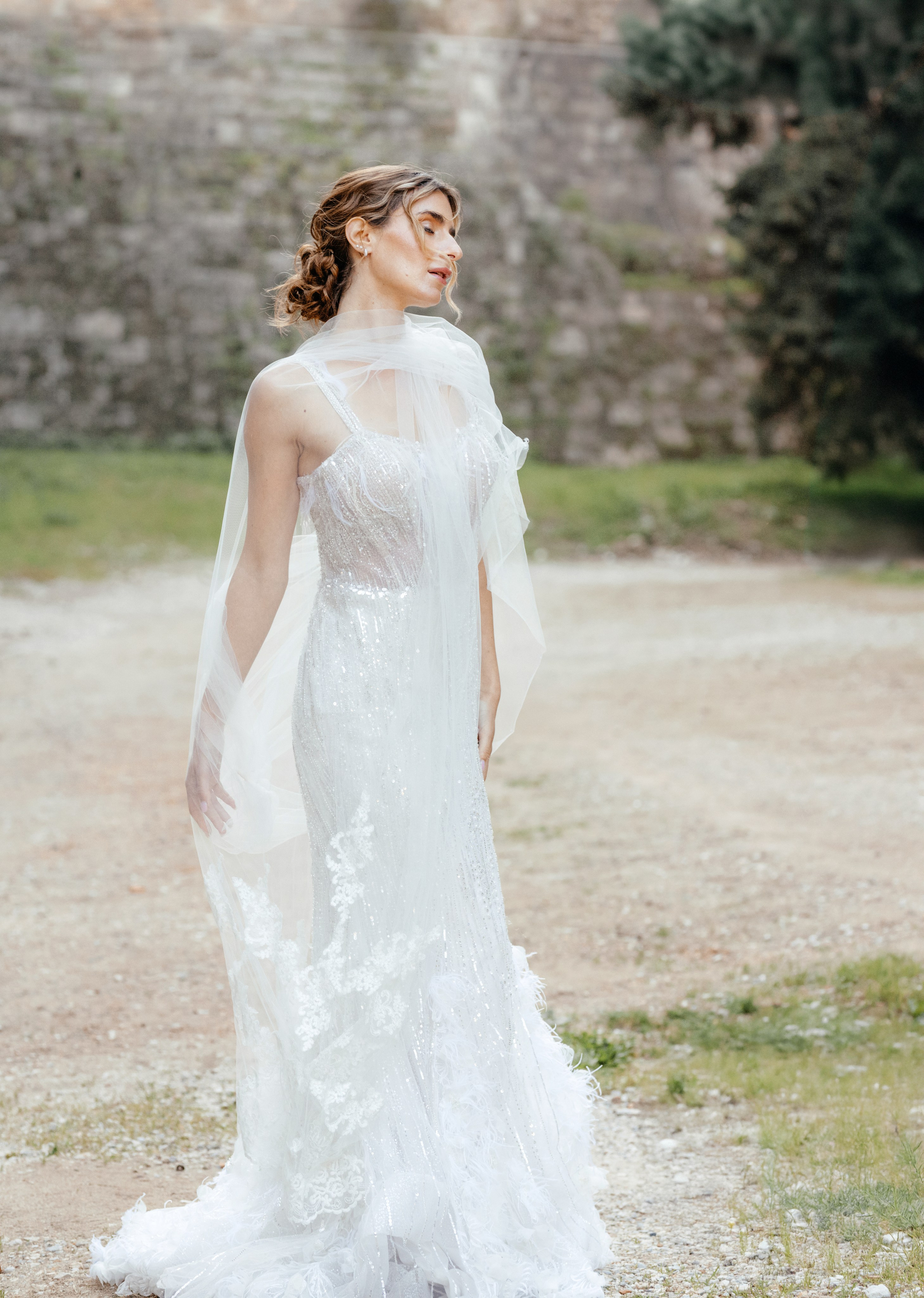 A radiant bride stands in the narrow, sunlit streets of Rhodes' Old Town, her intricate lace wedding dress contrasting beautifully with the weathered stone walls behind her. The editorial-style portrait highlights her serene expression and the timeless elegance of the historic setting, with soft natural light accentuating her features.