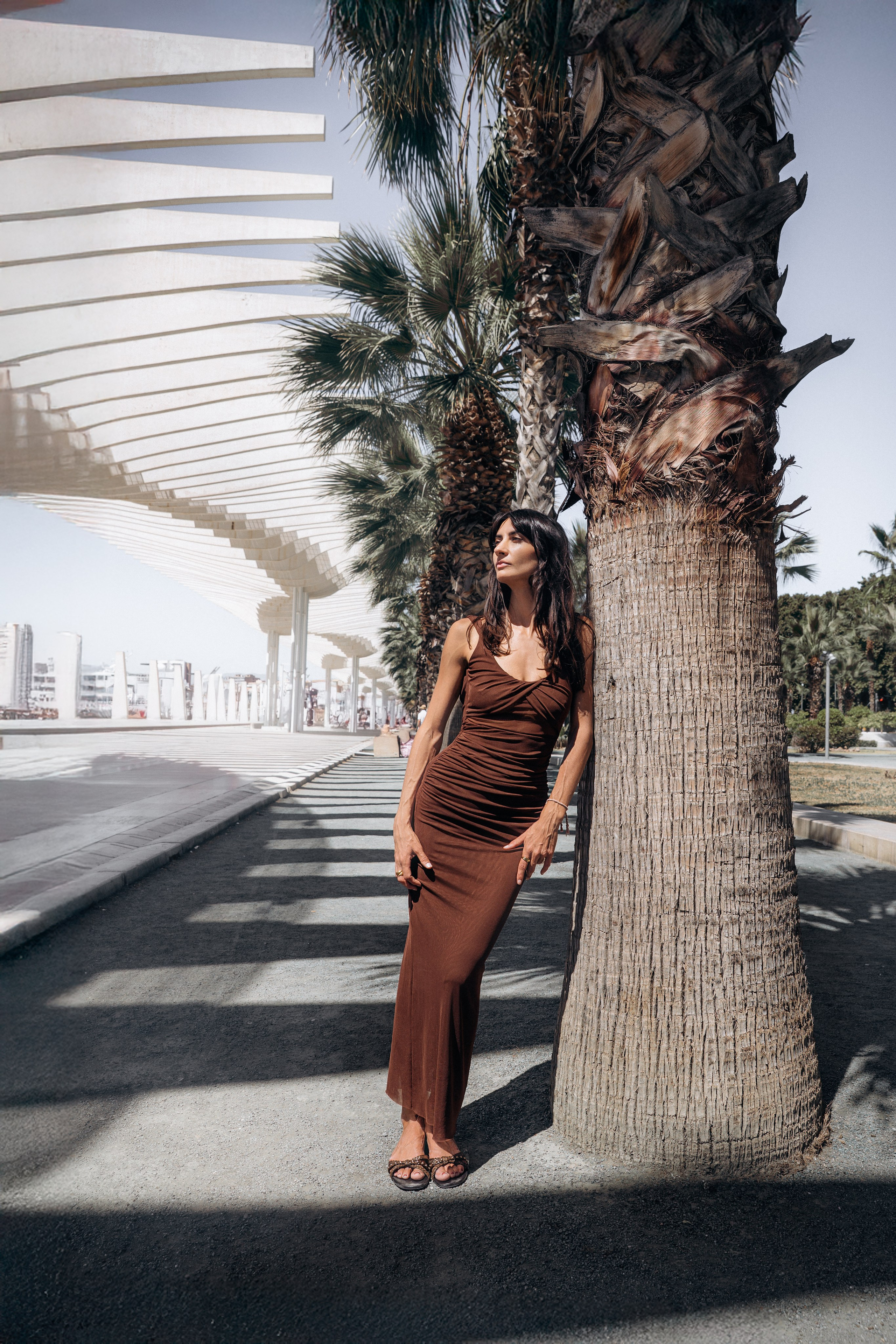 Стильная женщина в коричневом платье, облокотившаяся на пальму вдоль современной набережной Muelle Uno в Малаге, Испания — модная уличная портретная фотография, сочетающая городской стиль, средиземноморские пальмы и теплое солнце, идеально подходит для портретных фотосессий в Малаге и Испании.
