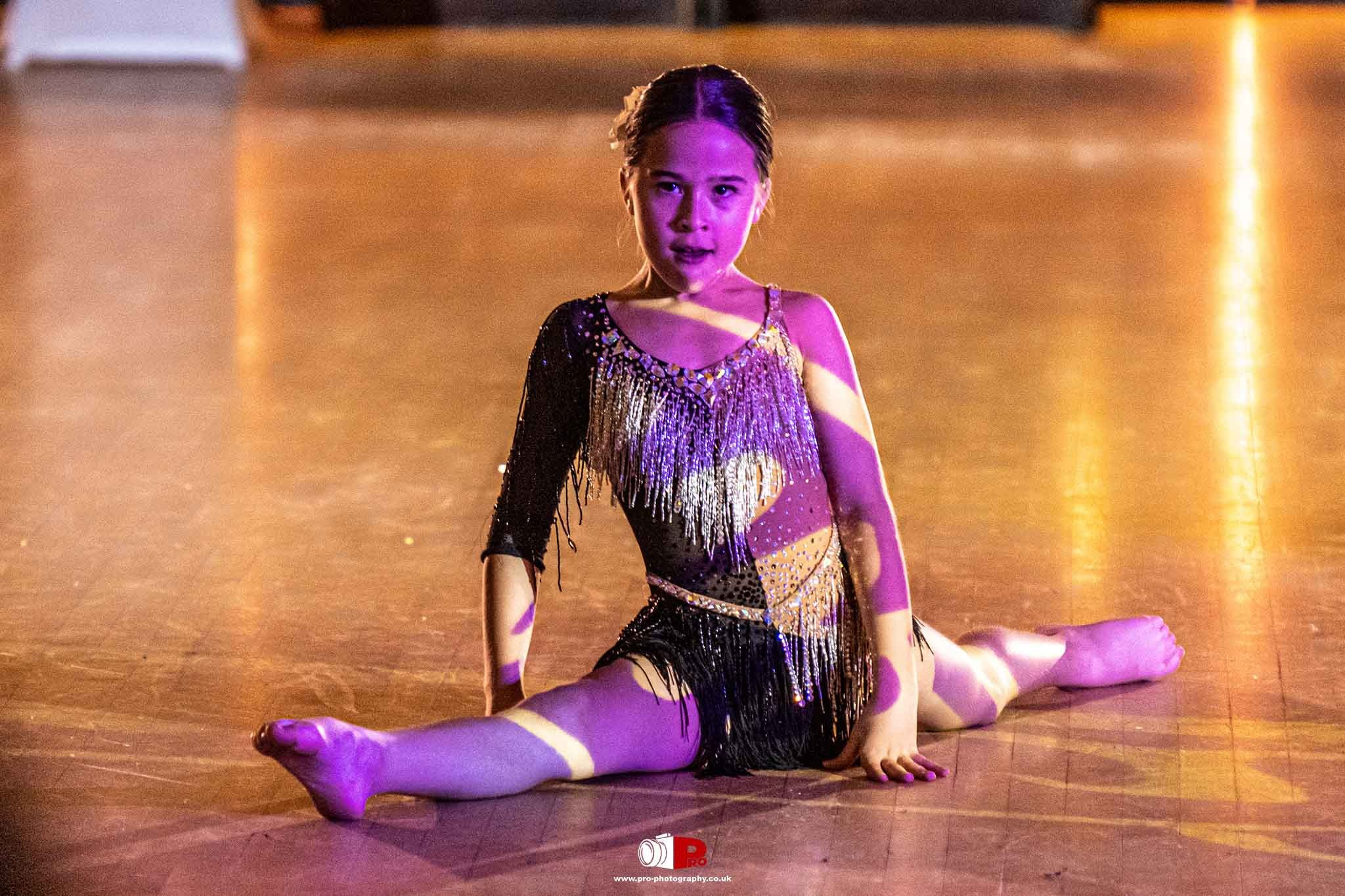 A young female dancer performing the splits in a sparkling black and gold outfit under dramatic stage lighting.