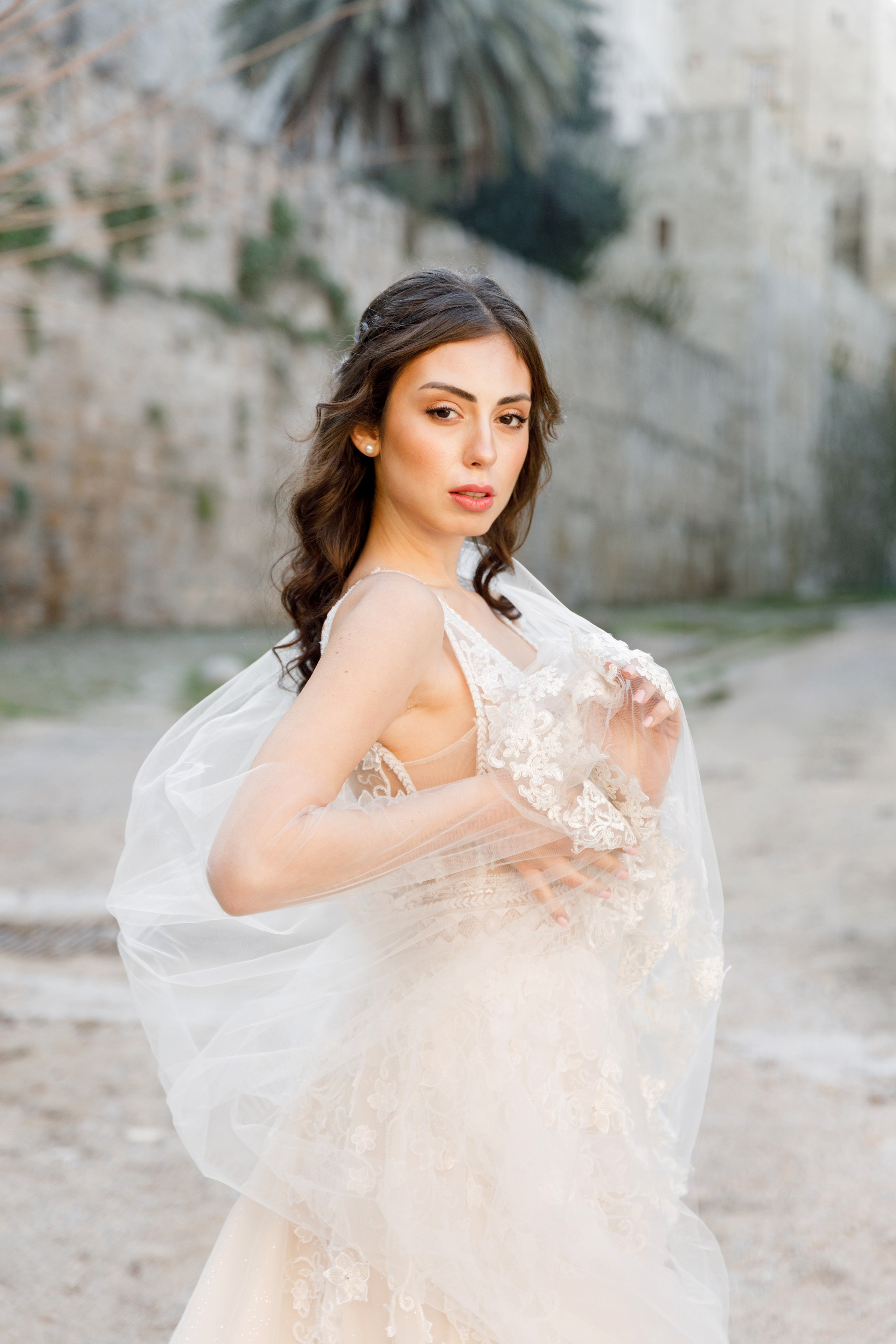 A stunning bride gazes thoughtfully in the enchanting alleys of Rhodes' Old Town, her flowing wedding dress complementing the rustic charm of the cobblestone streets and ancient architecture. The editorial-style portrait captures her poise and the romantic atmosphere of the medieval surroundings, bathed in warm, golden light.