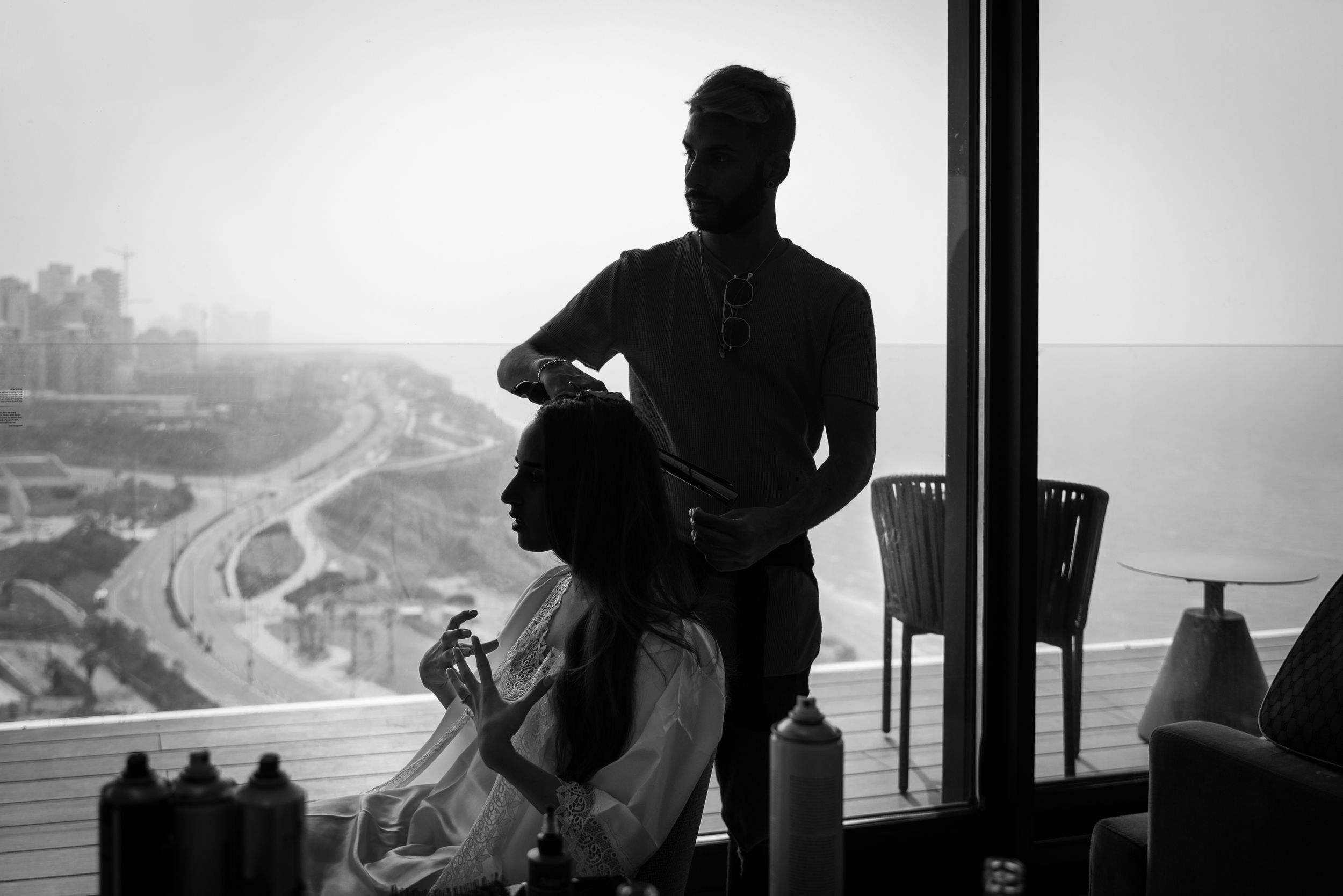 A hairstylist works on a bride’s hair beside a panoramic window overlooking the sea