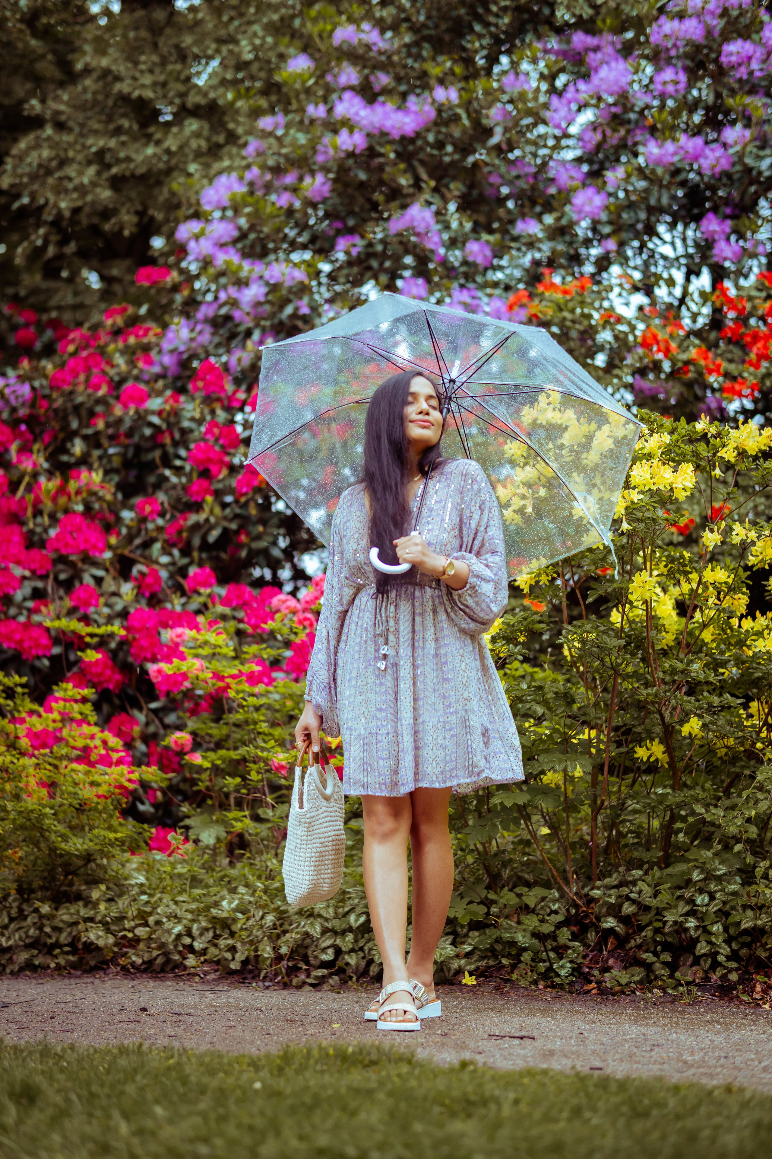 woman standing in a flower park and holding a transparent umbrella and a beach bag