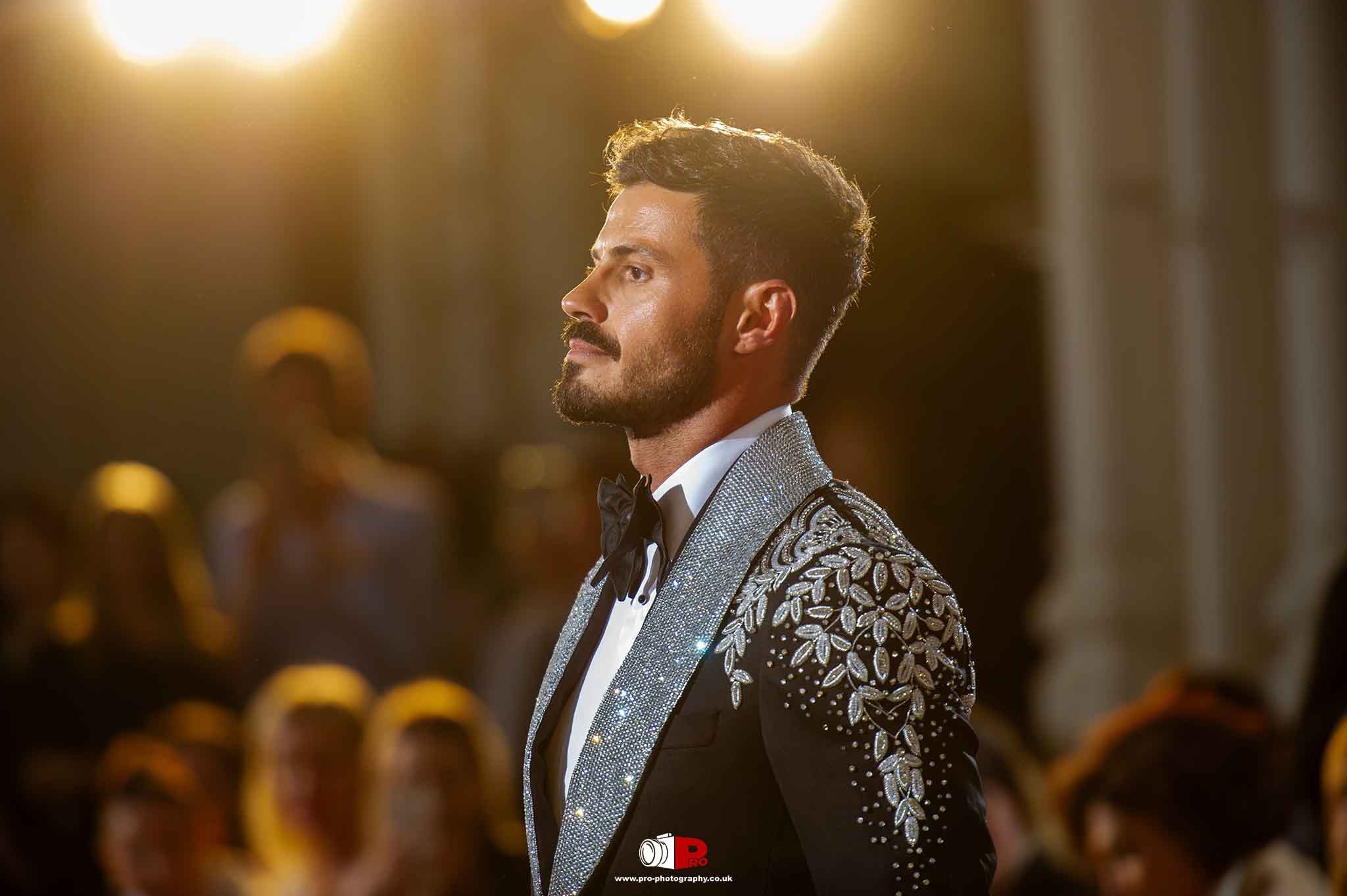 A male model walks down the runway in a dazzling silver-embellished tuxedo under spotlight lighting.