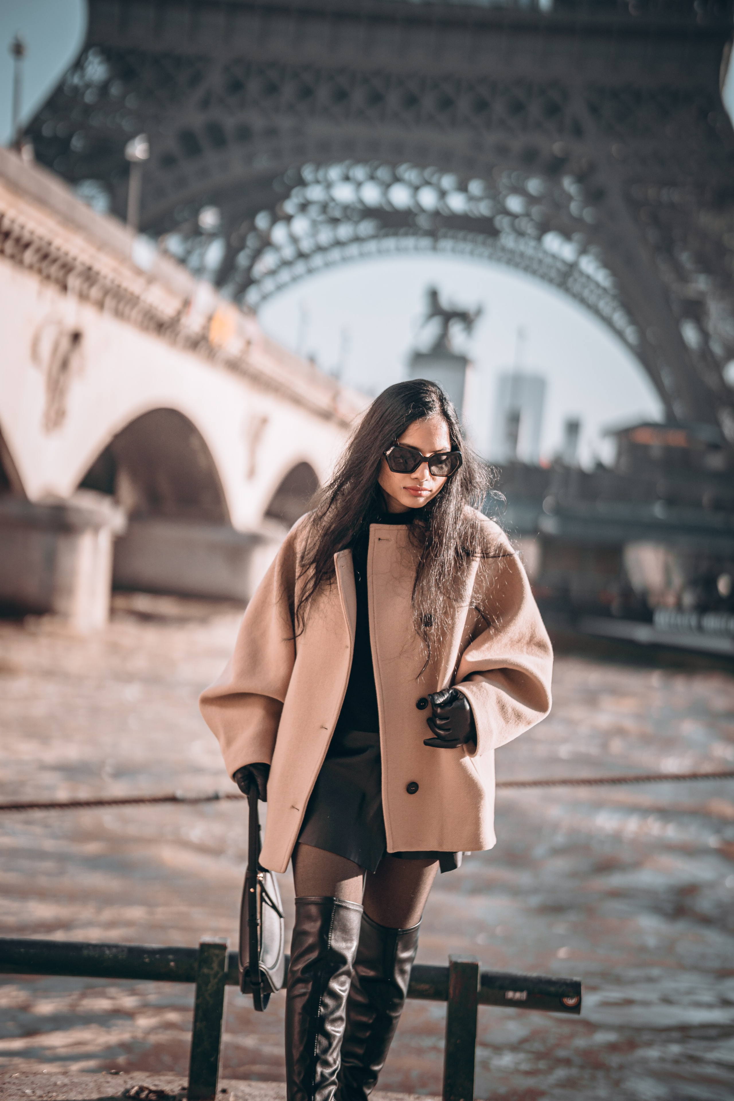 a woman wearing oversized brown jacket and standing with paris bridge and eiffel tower view behind her
