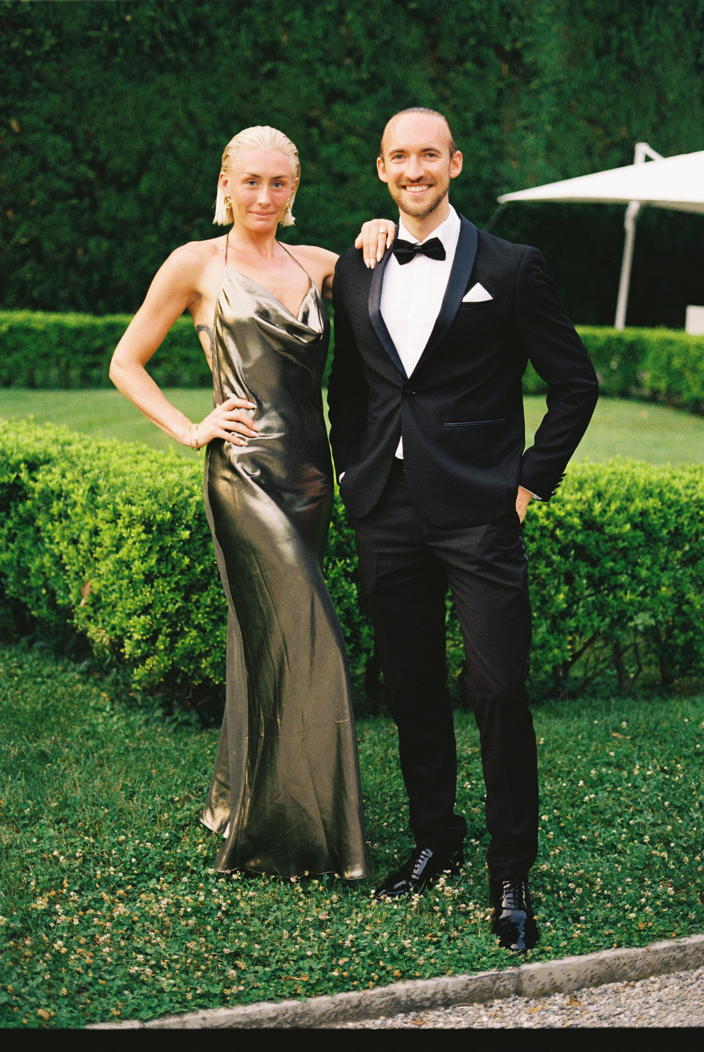 Couple in formal attire poses confidently in garden, with woman in green metallic dress.