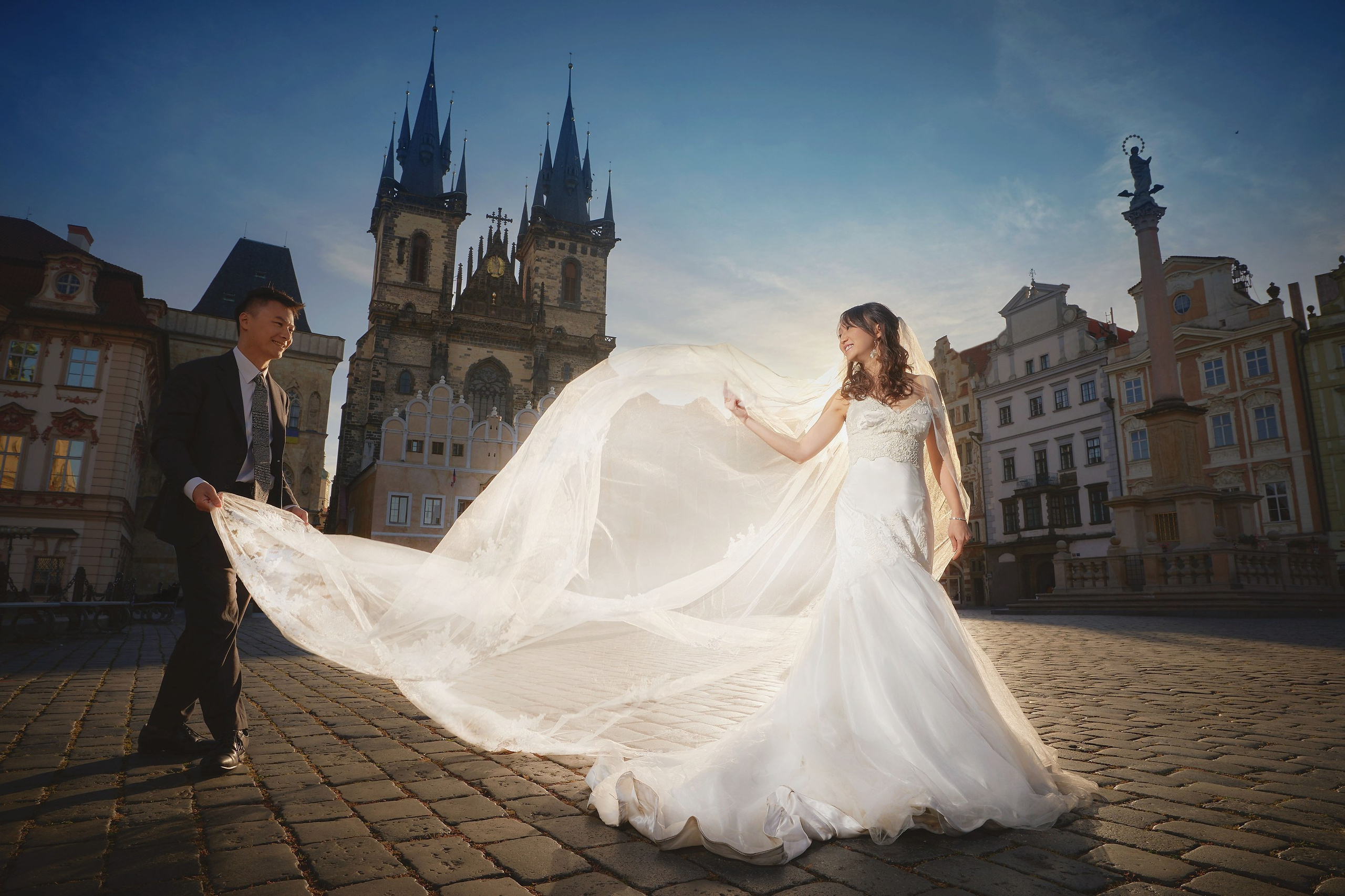 Asian bride's veil is lifted by her groom as the sun backlights the scene in the heart of the Old Town Square in Prague shortly after sunrise
