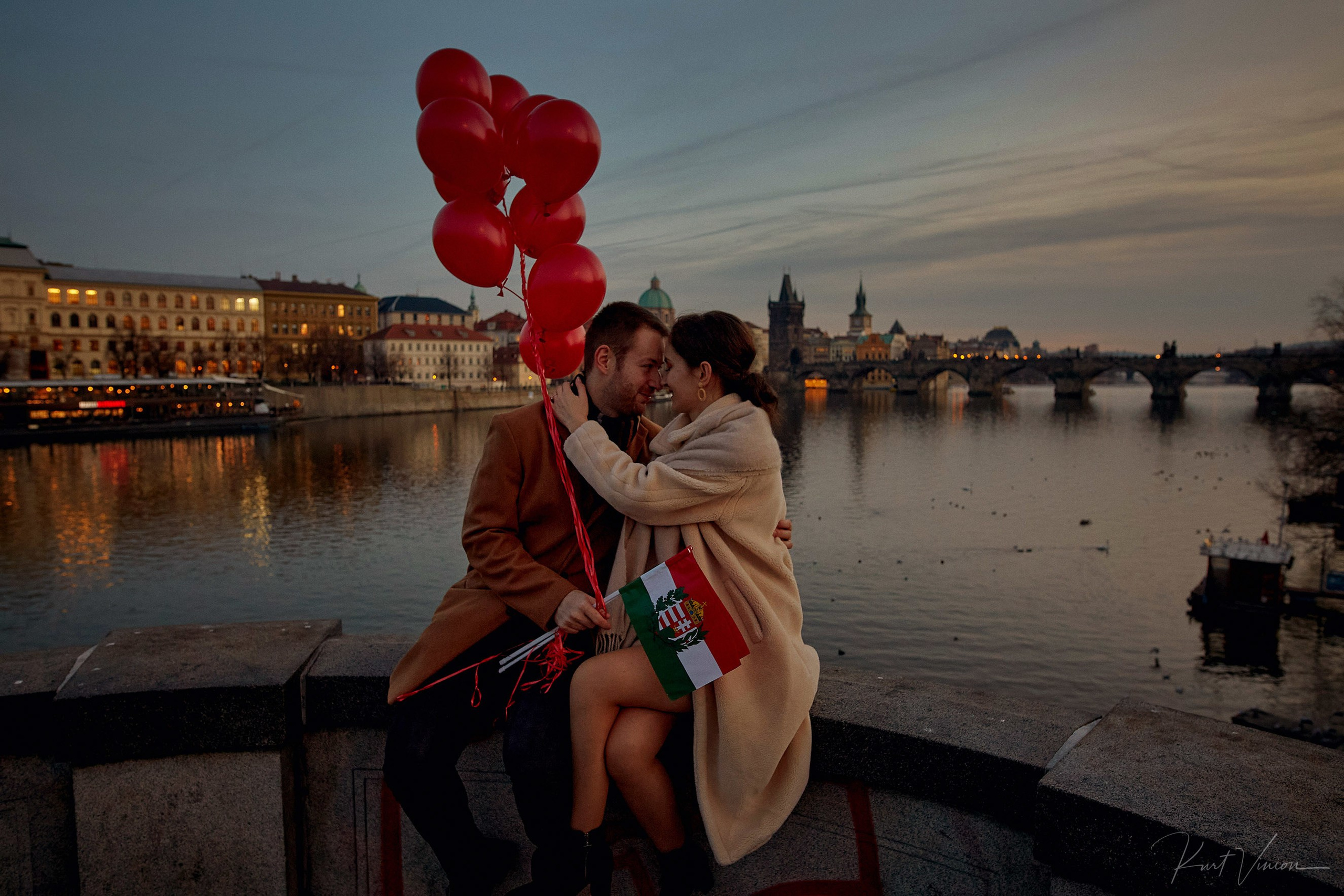 Bulgarian couple embracing with red balloons and flag above river by Charles Bridge in Prague.