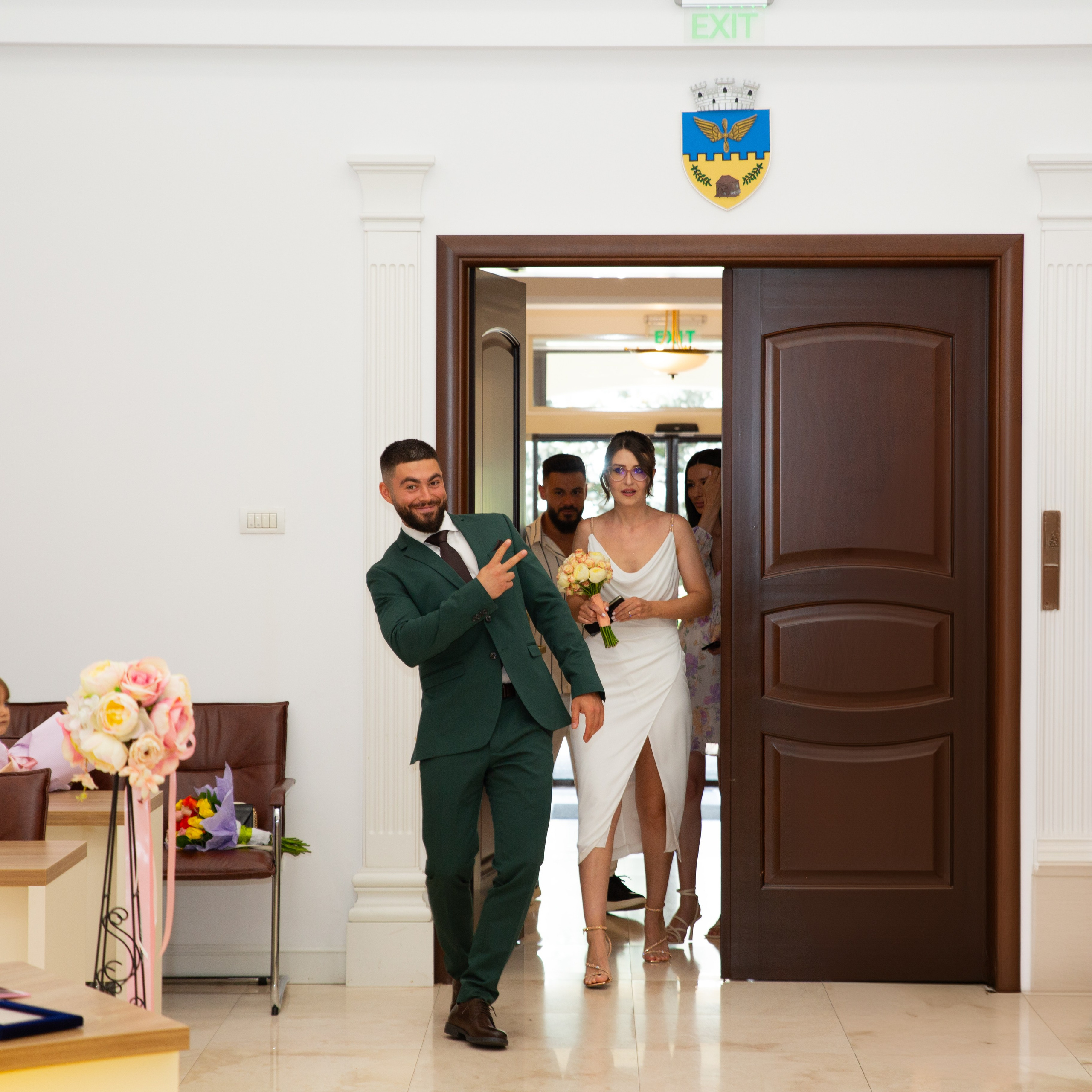Bride in white dress holding bouquet and groom in dark suit walking through glass doors.