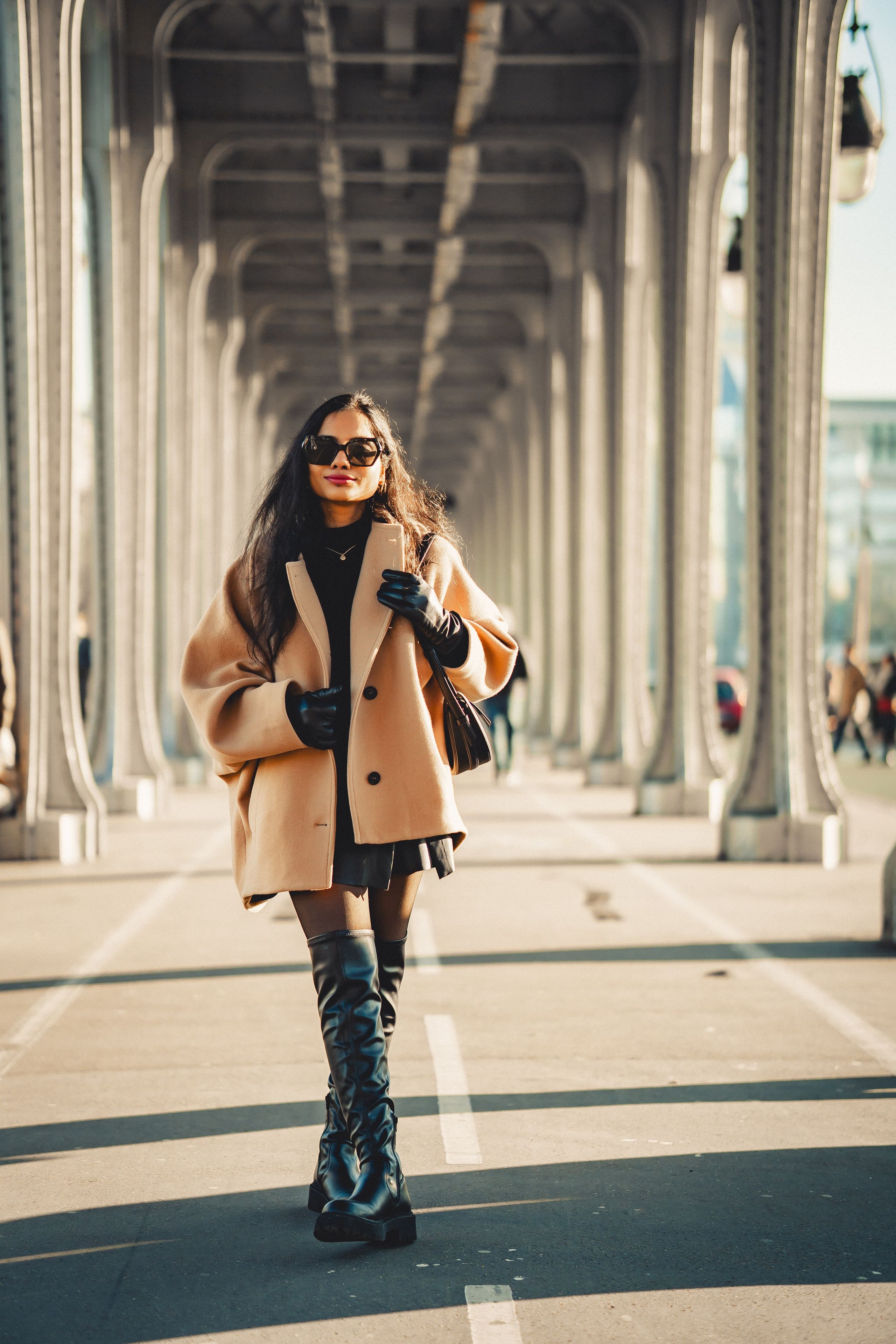 a woman wearing oversized brown jacket and knee high black boots and sunglasses and standing at bir hakim bridge